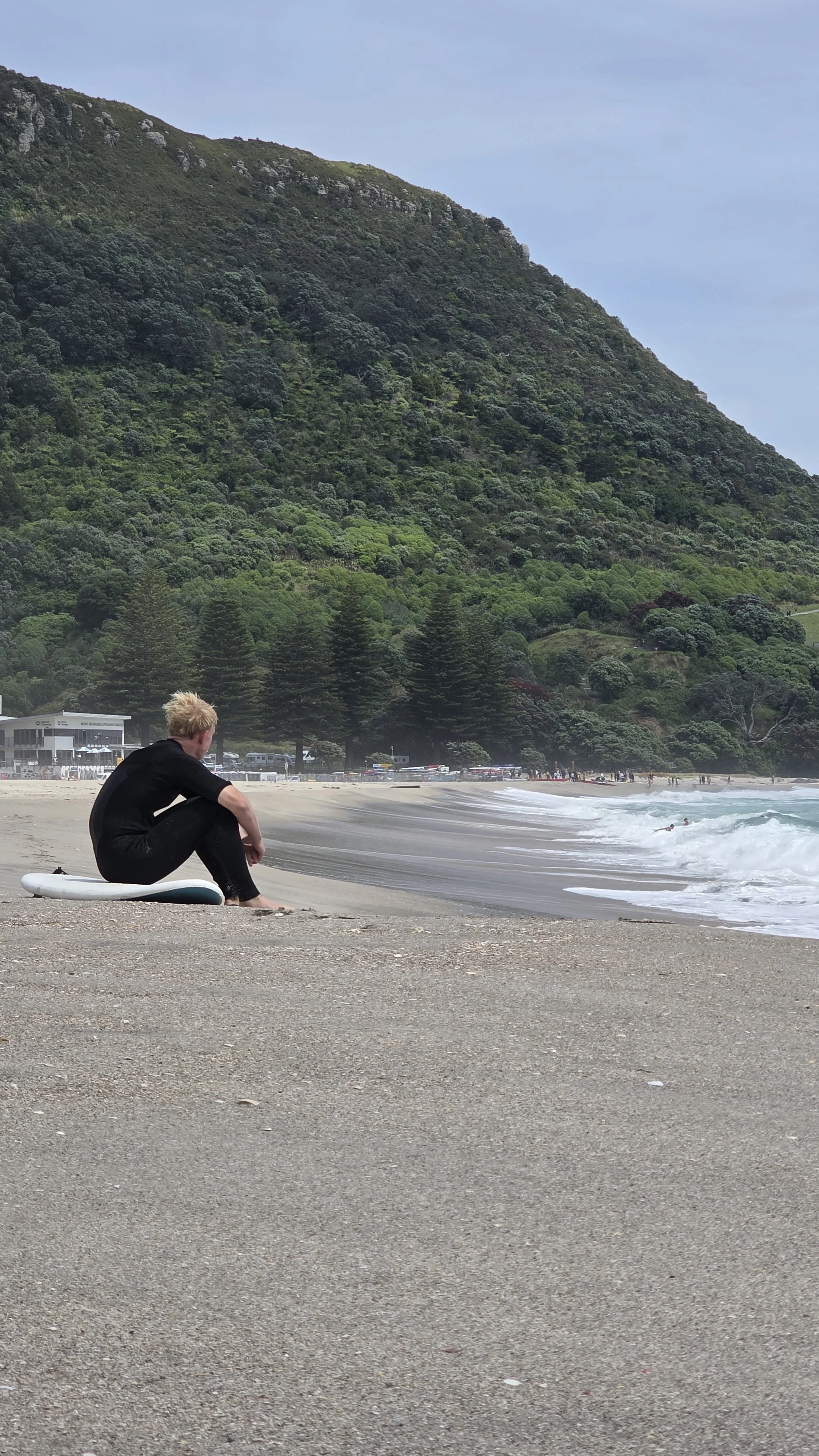 A person sitting on a surfboard on a sandy beach, facing the ocean with waves, green hills, and trees in the background.