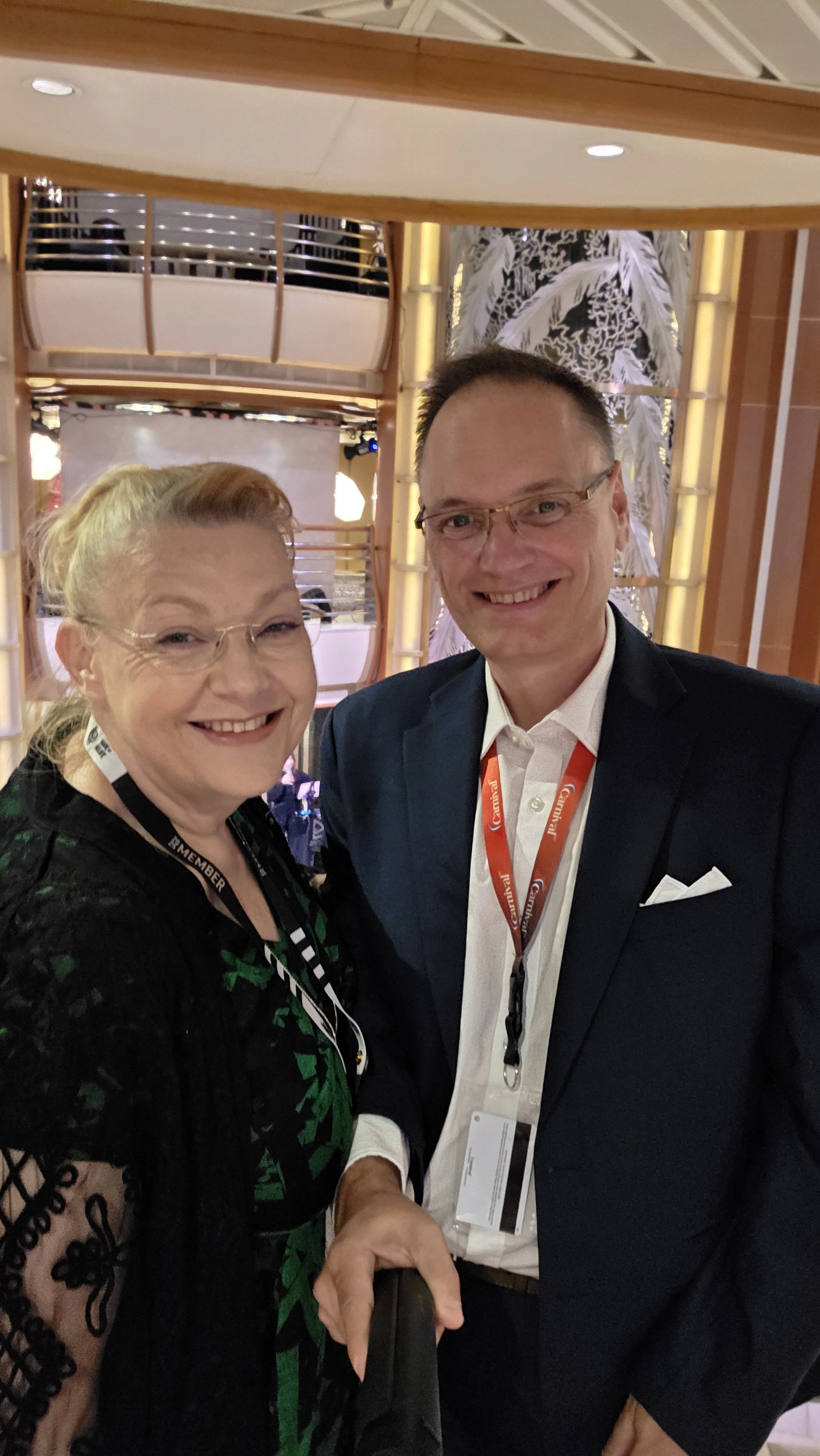 Two people taking a selfie at a conference or event, smiling and wearing badges around their necks, with a decorative background in a well-lit indoor setting.
