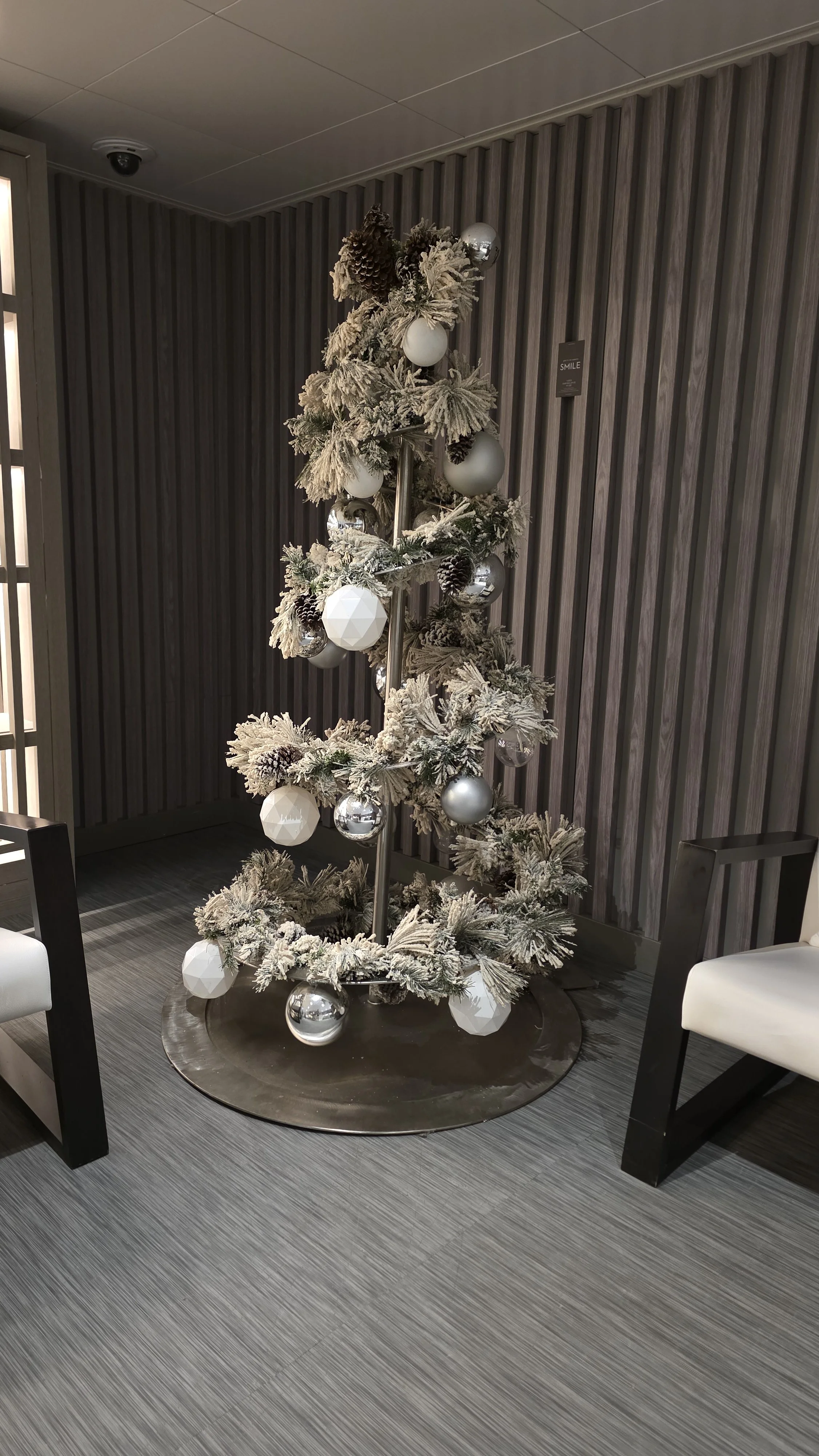 A Christmas tree decorated with white, silver, and gray ornaments and frosted artificial snow. The tree is placed in a modern indoor setting with gray textured wall panels and gray carpeting, flanked by white and black seating.