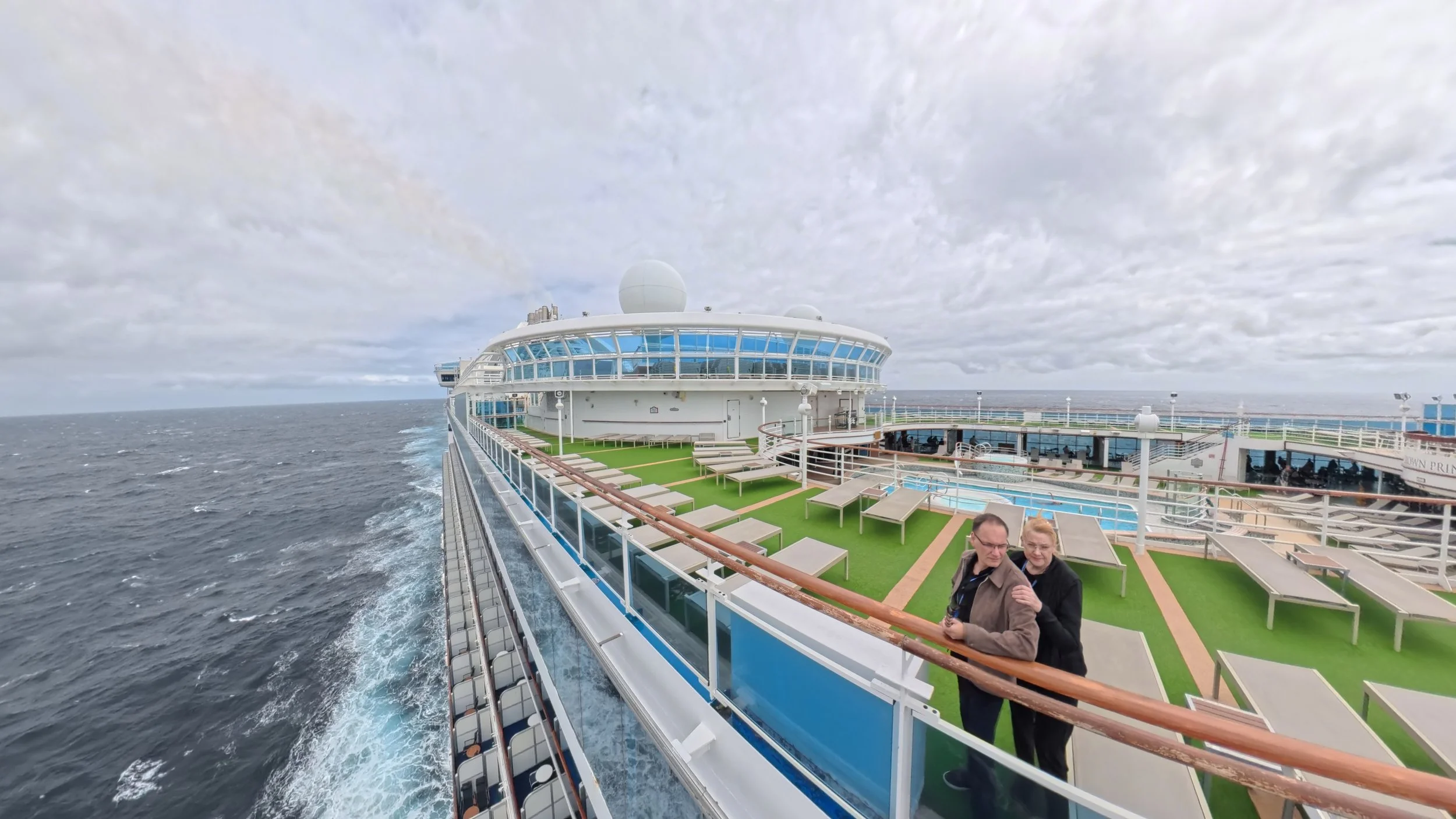 Two people standing on the deck of a large cruise ship, with lounge chairs, a small pool, and ocean in the background under a cloudy sky.