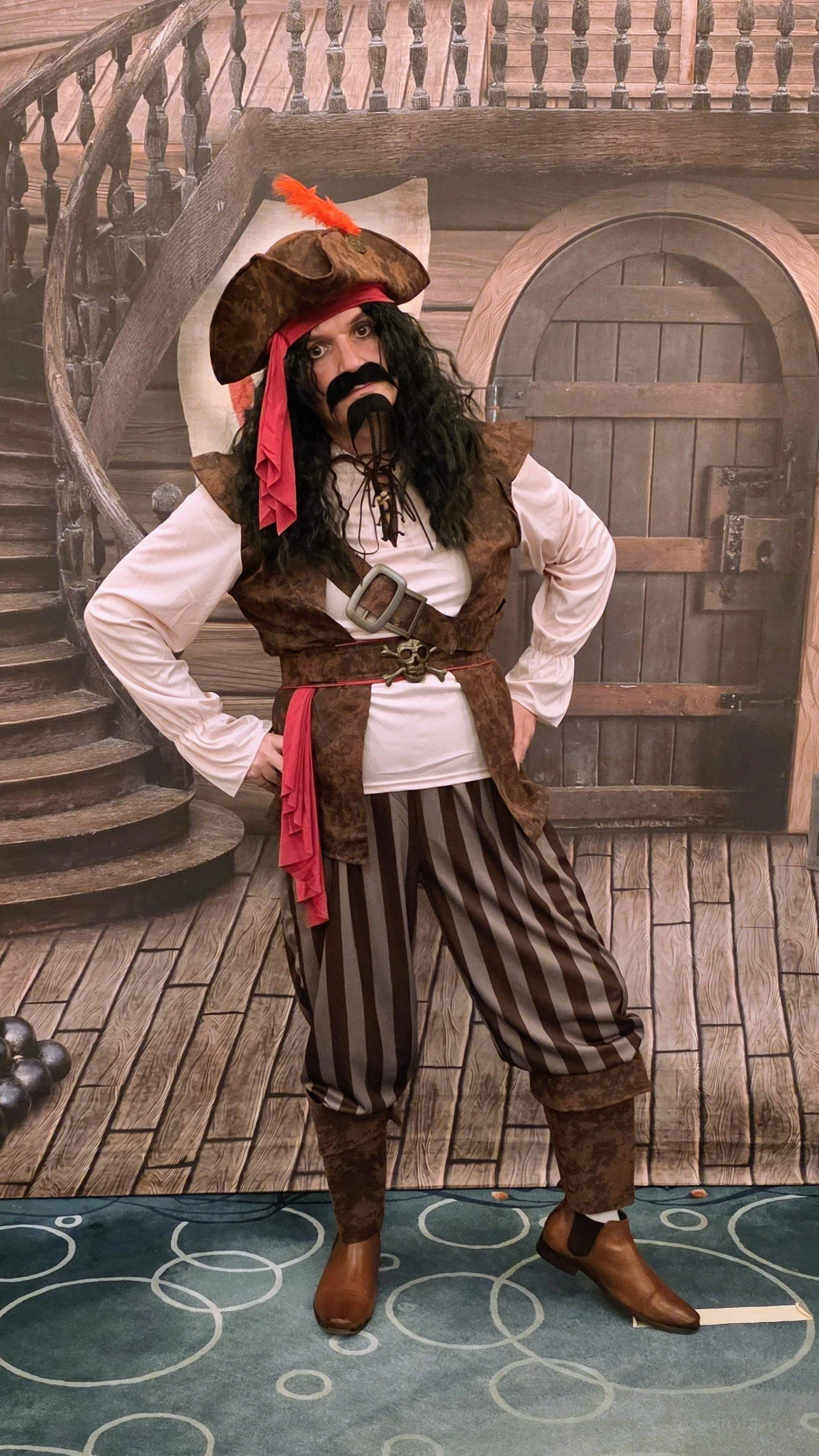 Person dressed as a pirate in front of a themed backdrop resembling a medieval door and staircase, with a faux wooden floor and a green carpet with white circles.