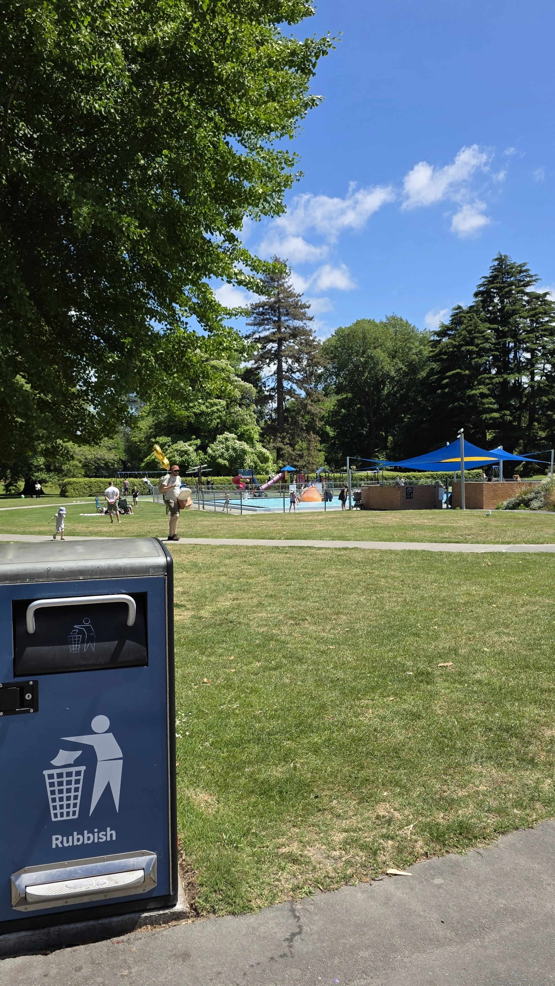 Scene of a local park with a swimming pool, slides, and people enjoying a sunny day.