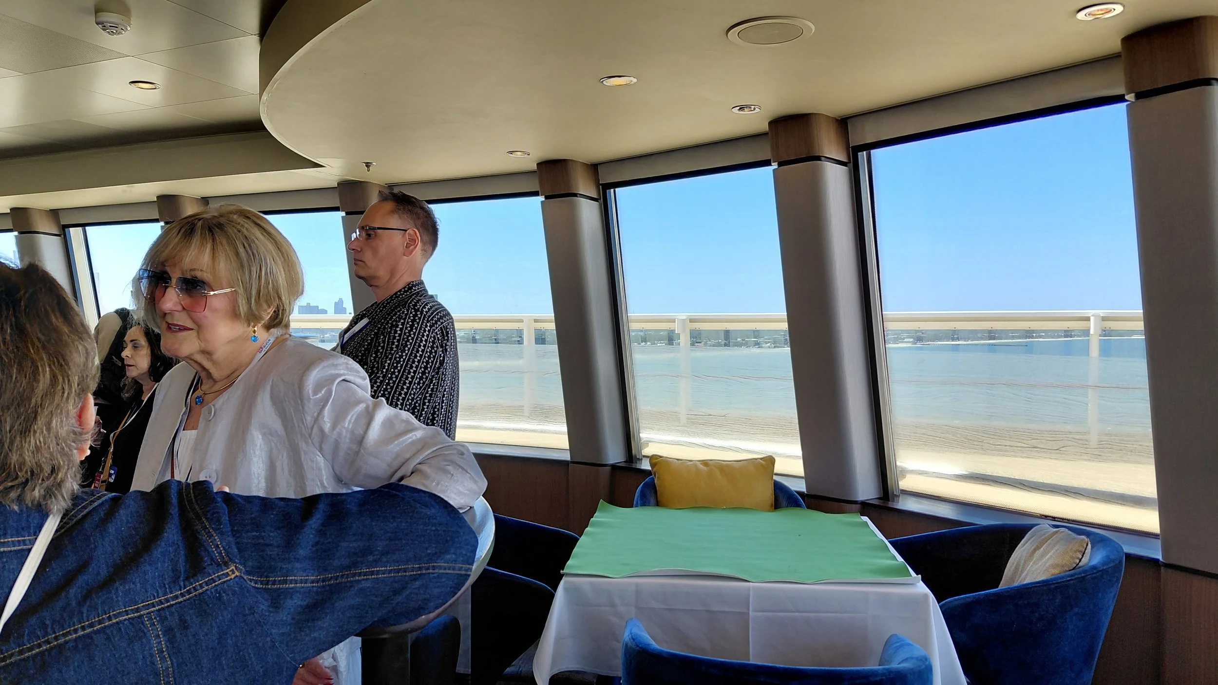 People socializing inside a restaurant with large windows overlooking a river or waterway on a sunny day.