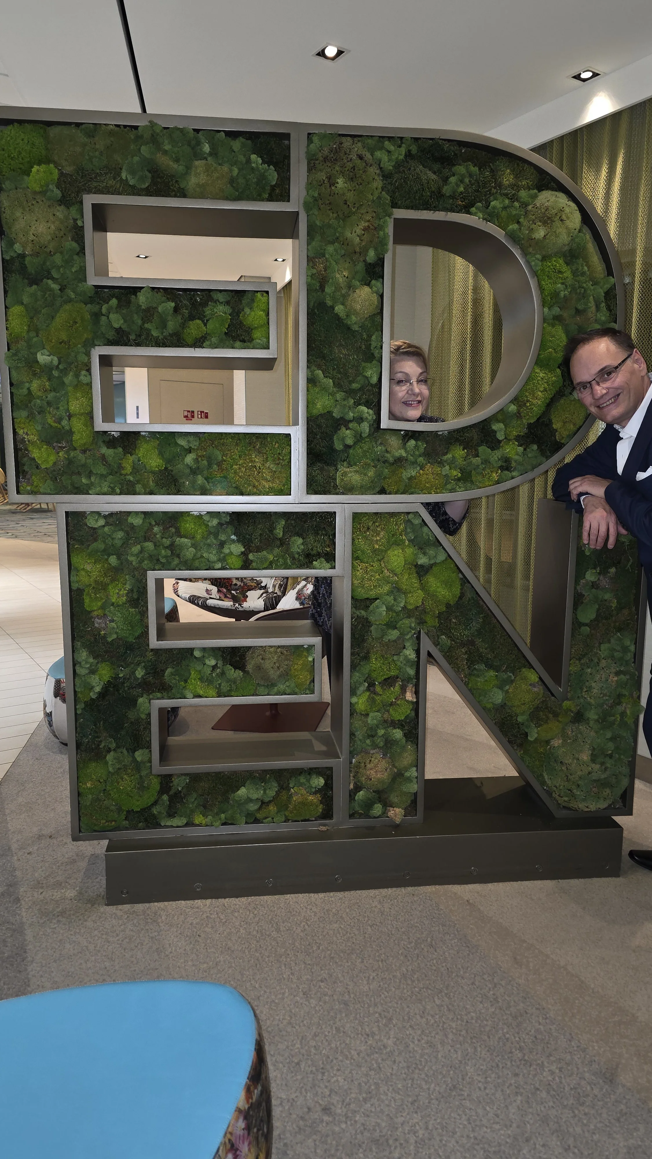 People peeking through a large decorative sign with moss-covered panels and metal framing.