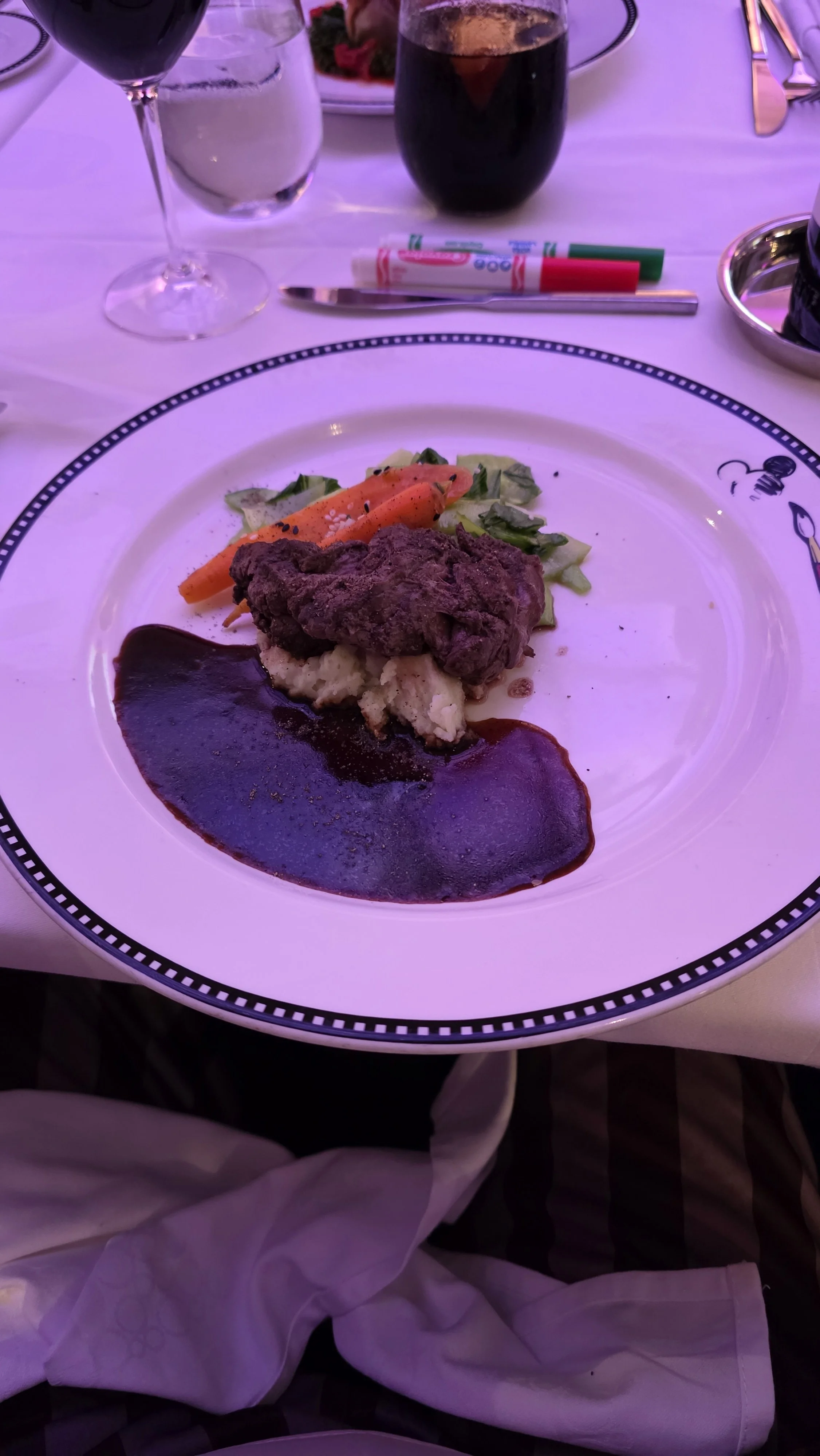 A plated meal featuring beef with sauce, mashed potatoes, and vegetables, set on a decorated dinner plate with drinks and utensils on a table.