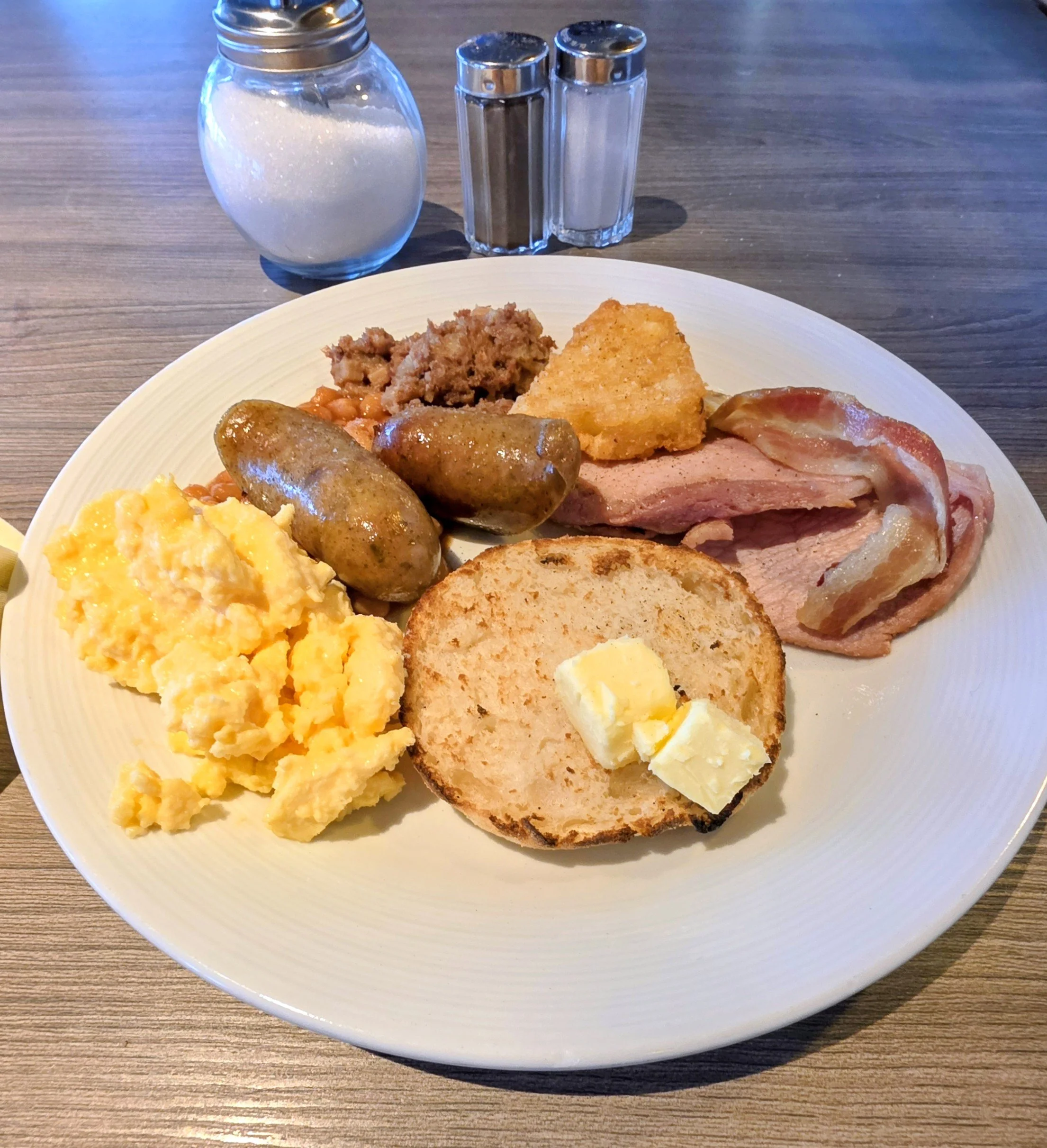 A plate of breakfast food including scrambled eggs, two sausages, a slice of toasted bread with butter, bacon, ham, a hash brown, baked beans, and corned beef hash, with salt and pepper shakers and a sugar dispenser in the background.