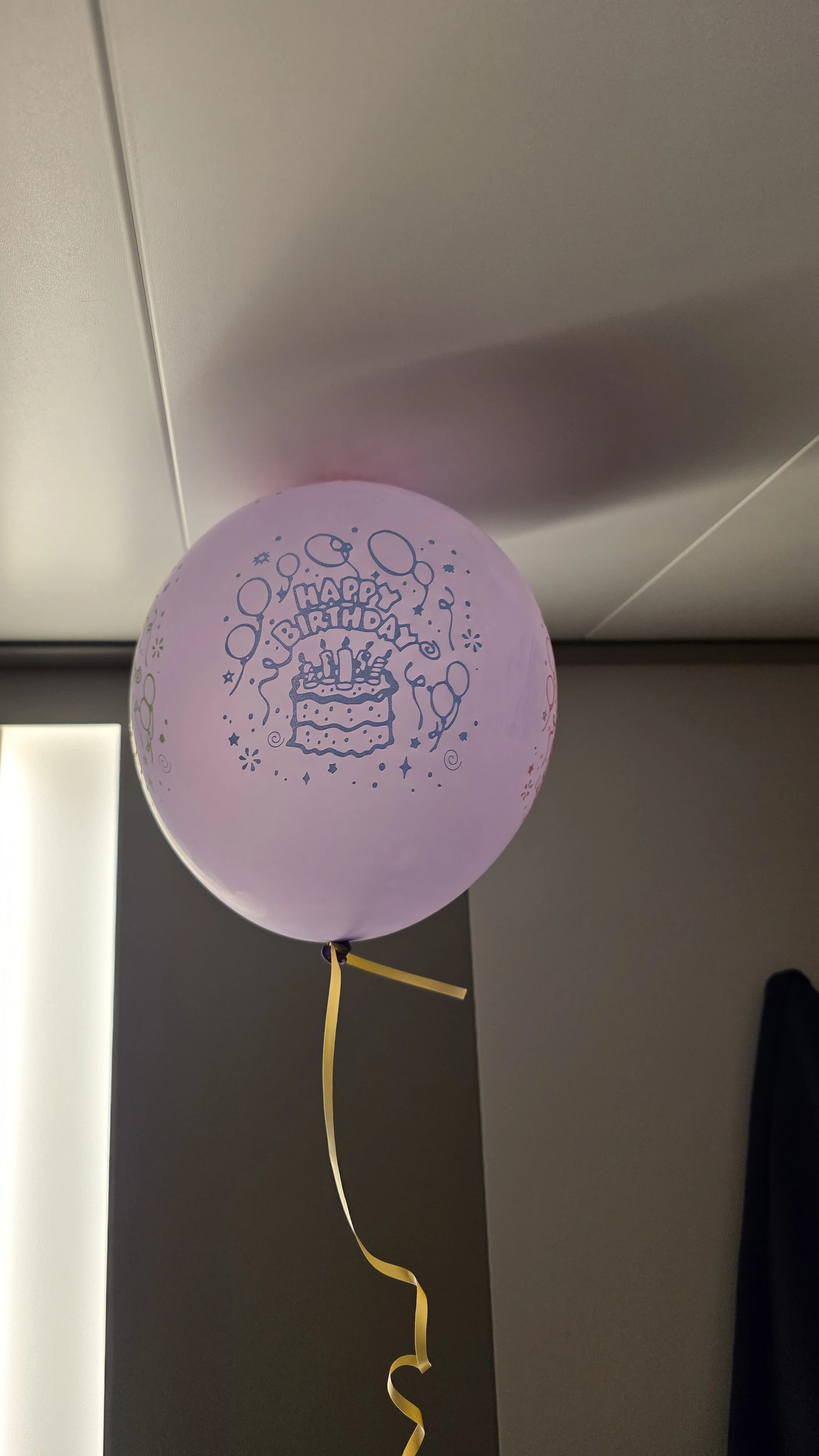 Pink birthday balloon with a birthday cake, candles, and 'Happy Birthday' written on it, floating indoors with a yellow ribbon.