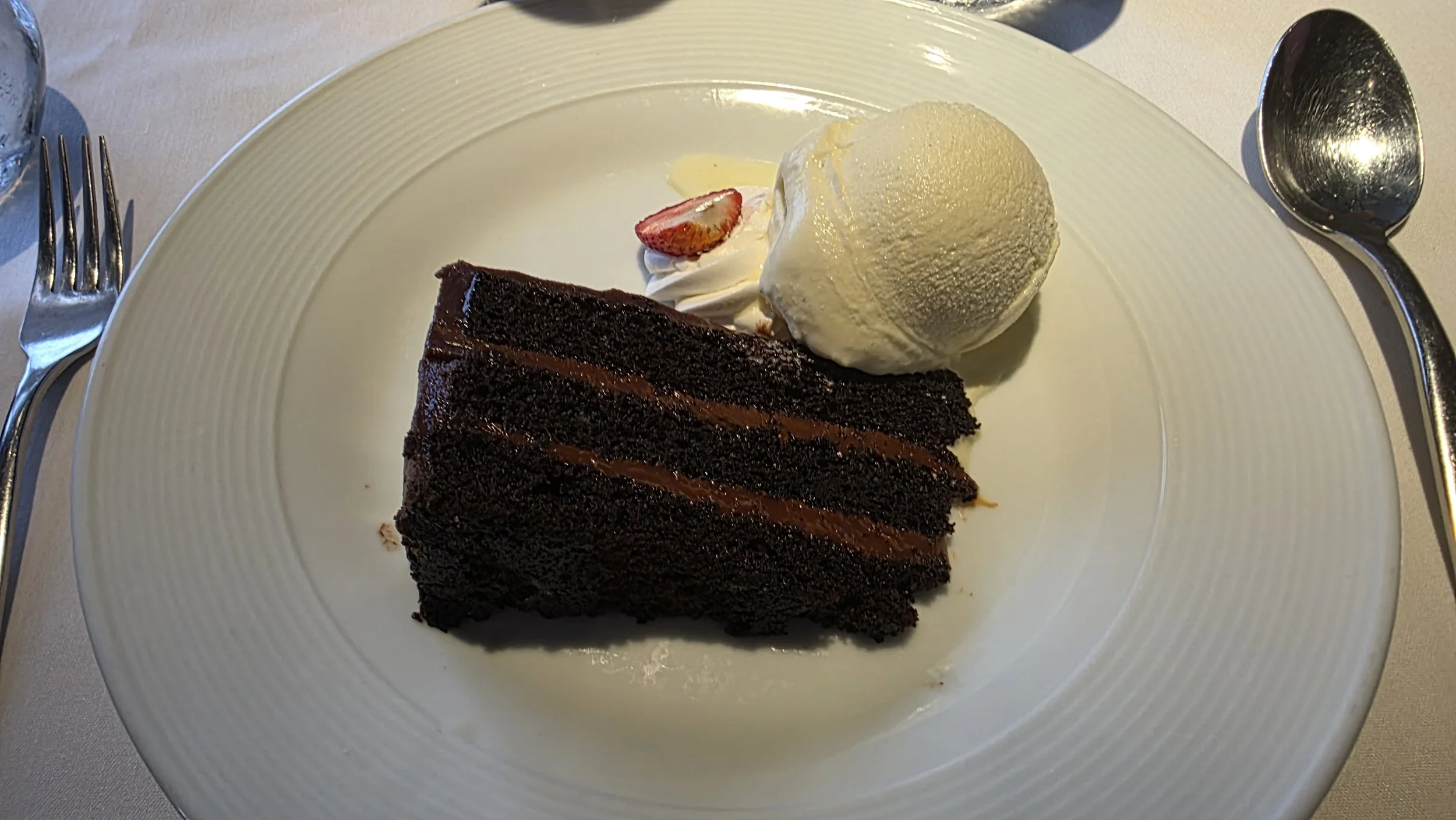 Slice of chocolate cake with layers of chocolate filling, accompanied by a scoop of vanilla ice cream and a dollop of whipped cream topped with a small strawberry slice on a white plate.