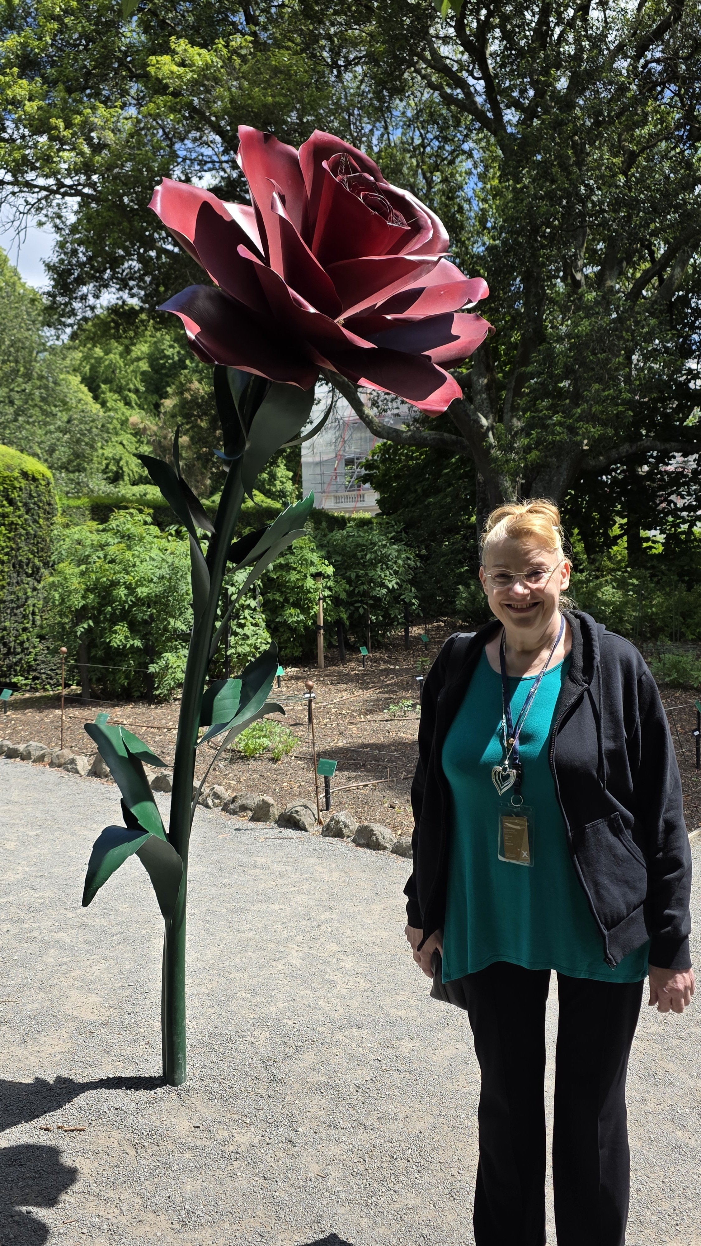 A woman standing next to a large artificial pink flower sculpture in a garden with trees and greenery, smiling and wearing glasses, a teal top, and a black jacket.
