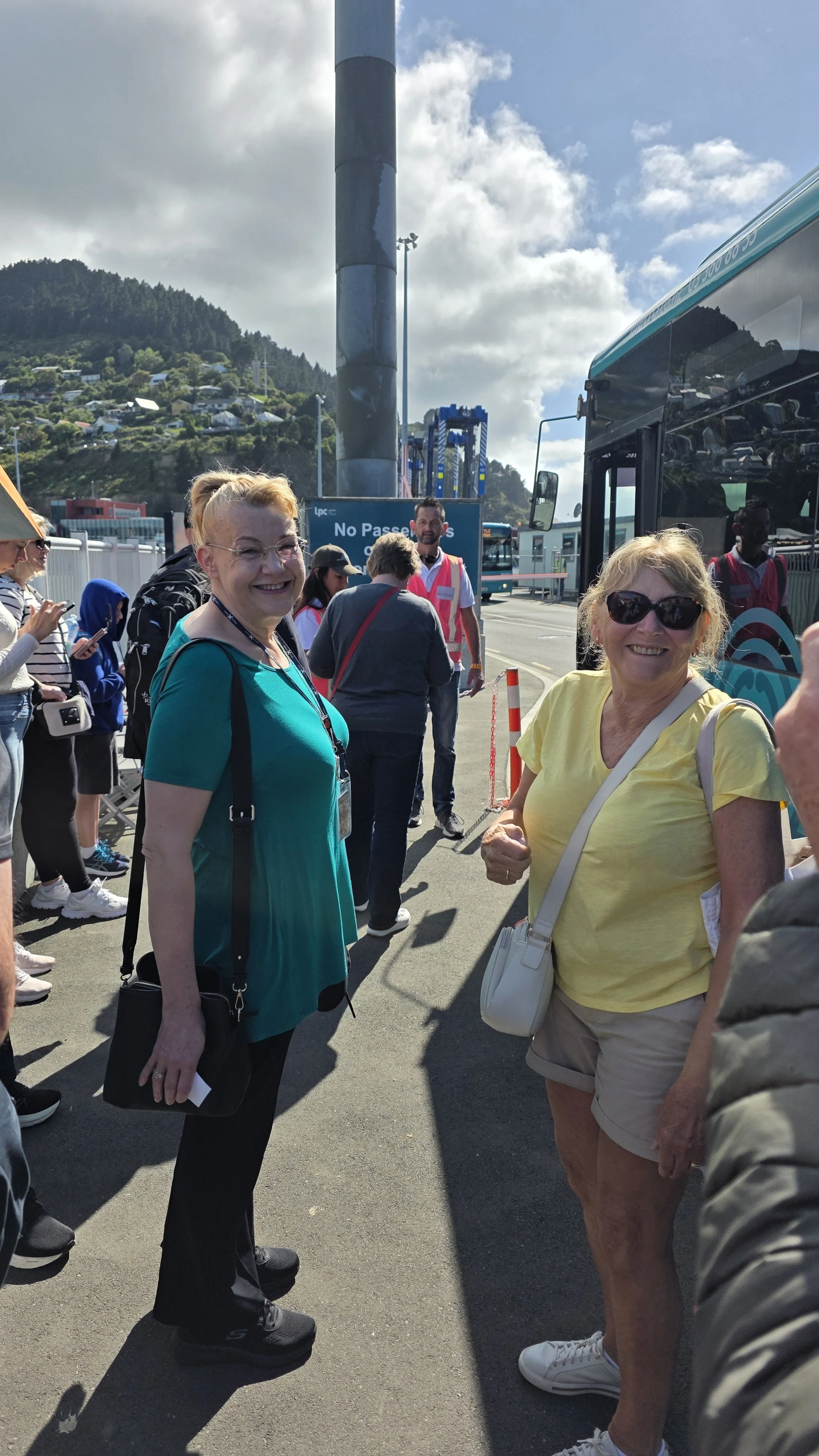 Two smiling women standing outdoors near a bus with a group of people in line behind them, with a hill and cloudy sky in the background.