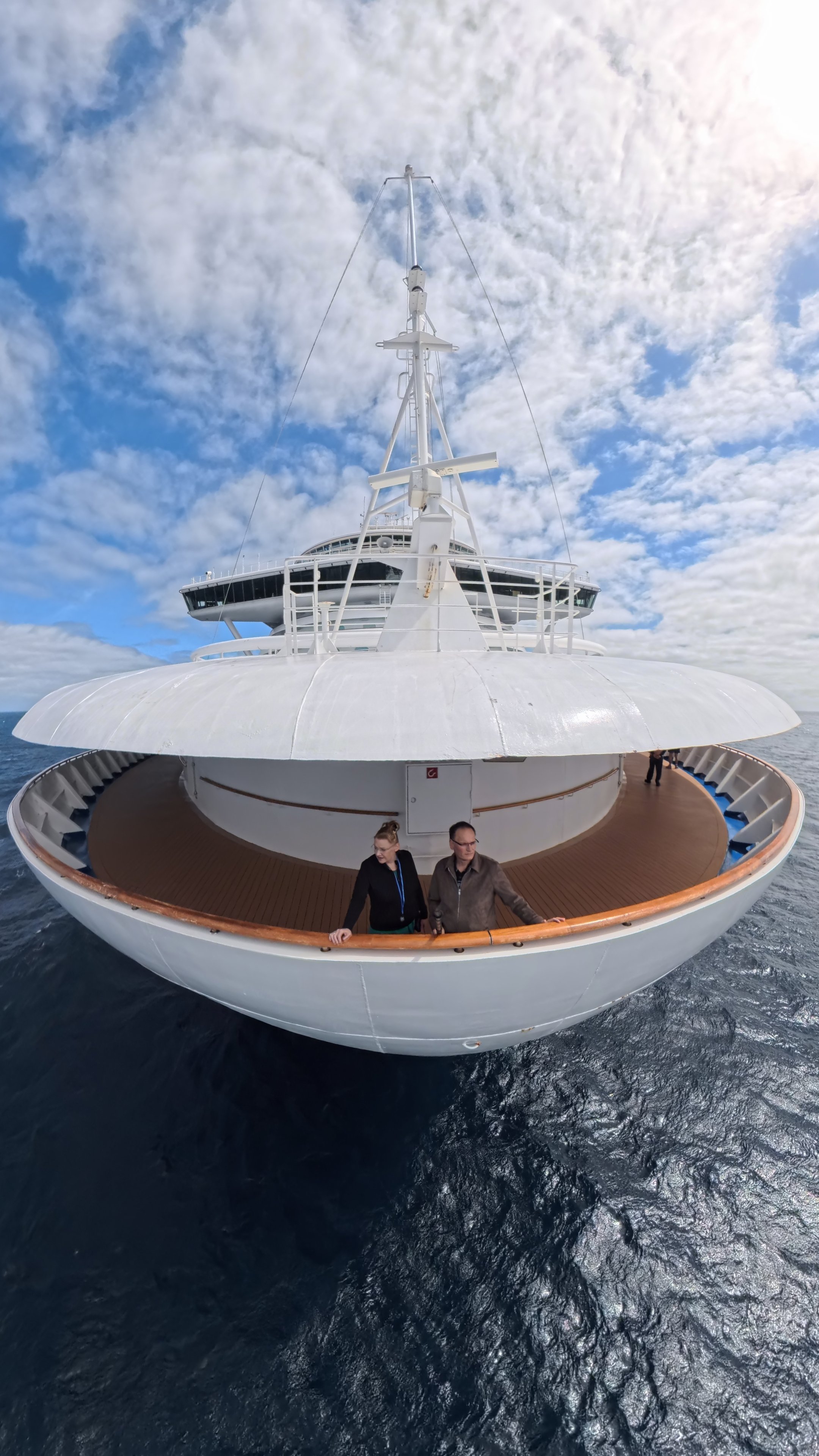 A large white yacht with a flat, circular deck and a central mast sails on the ocean under a partly cloudy sky. Two people are standing on the deck.
