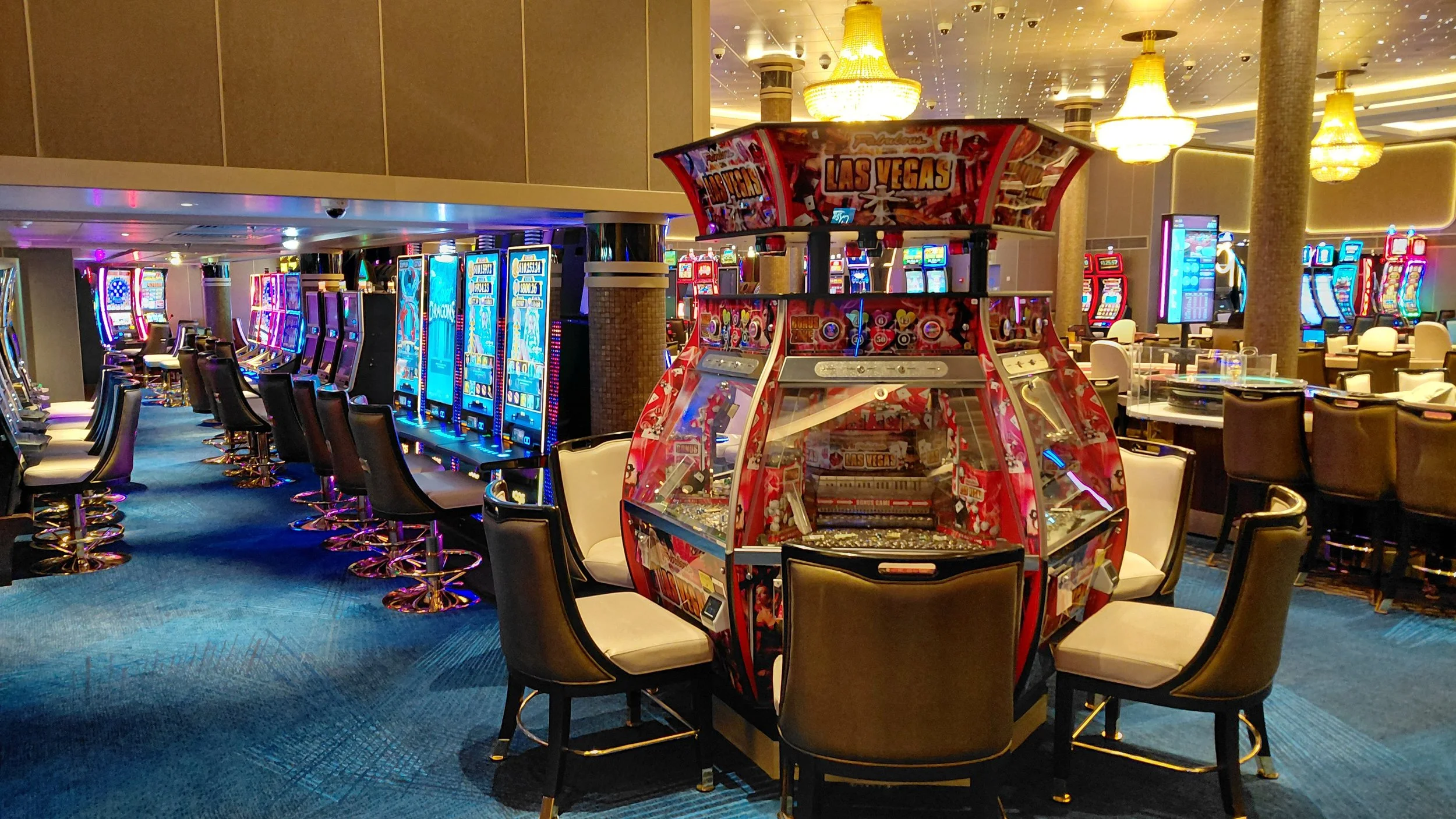 Inside a casino with rows of electronic slot machines and a central arcade game labeled 'Las Vegas,' surrounded by chairs and illuminated by chandeliers.