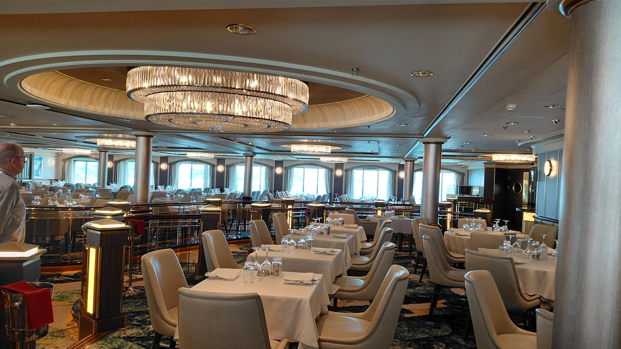 Elegant restaurant with round tables covered in white tablecloths, set with glasses and utensils, beige chairs, large windows letting in natural light, and modern chandeliers hanging from the ceiling.