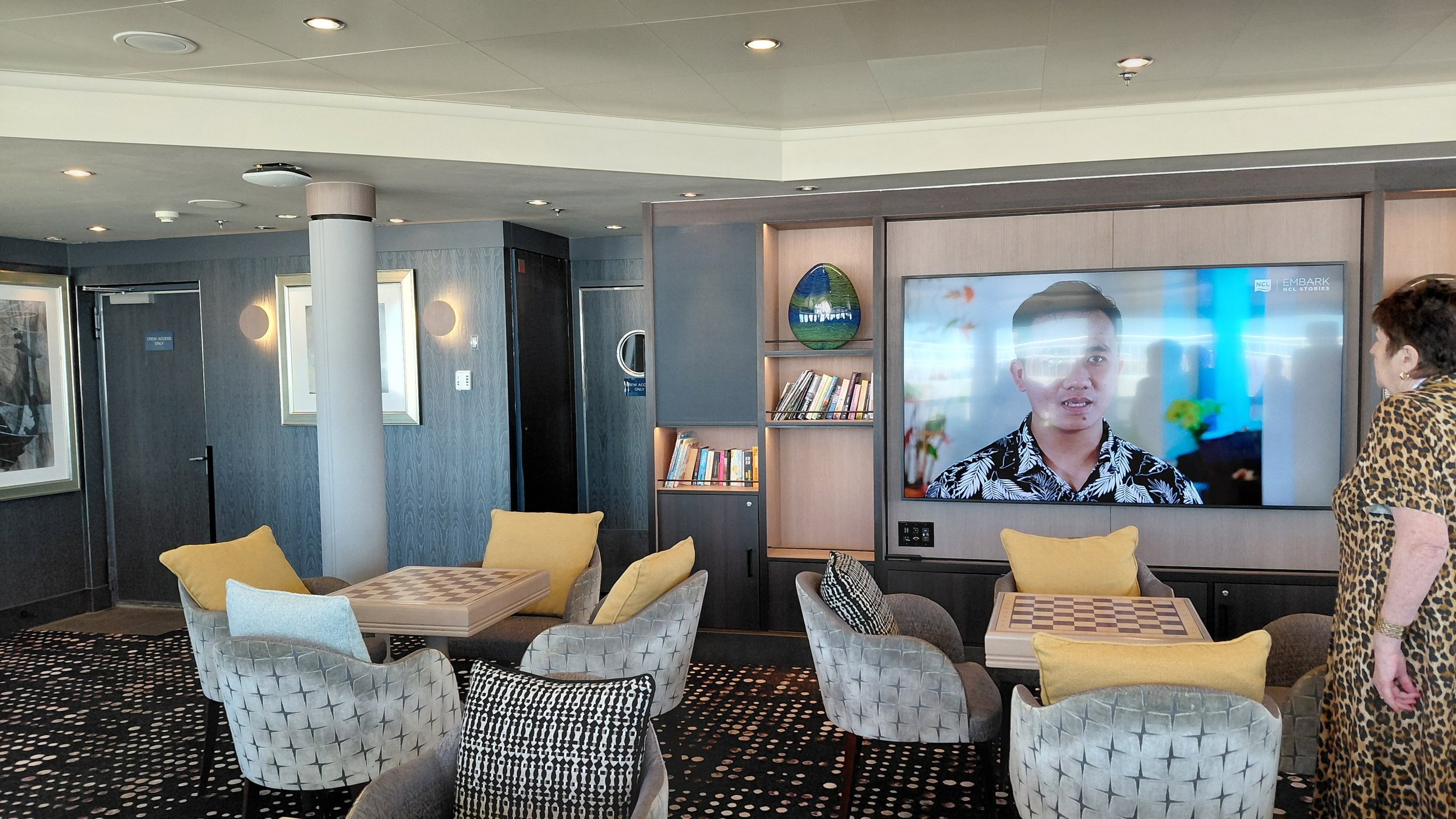A woman wearing a leopard print dress standing in a modern lounge area with a large wall-mounted television, bookshelves, and armchairs with yellow and gray pillows. The television displays a man in a floral shirt speaking.