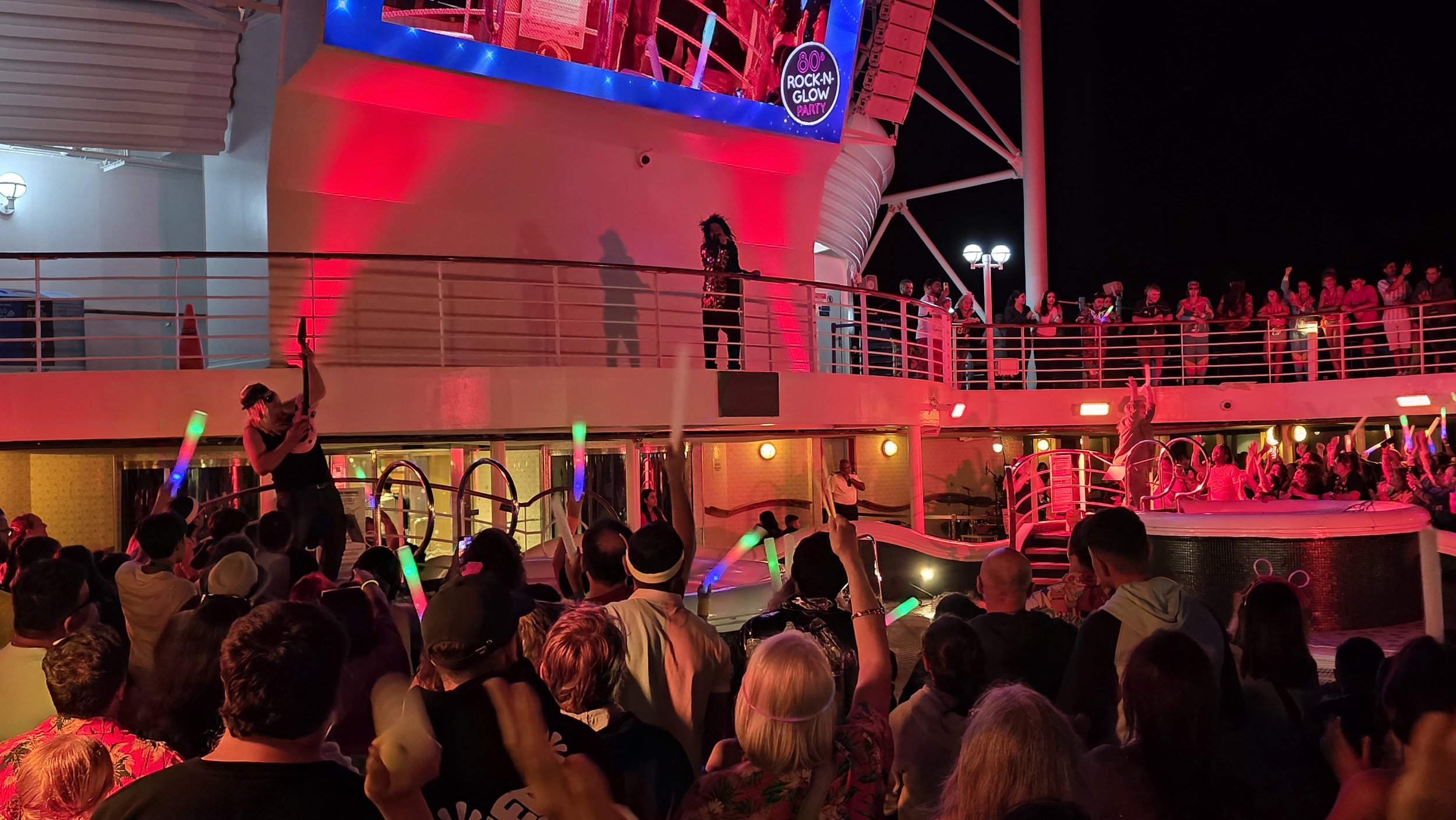 Nighttime scene of a crowded outdoor party deck with colorful lighting, people dancing and enjoying live entertainment on a boat or cruise ship, with a performer singing on a raised platform and a woman with a microphone in the background.