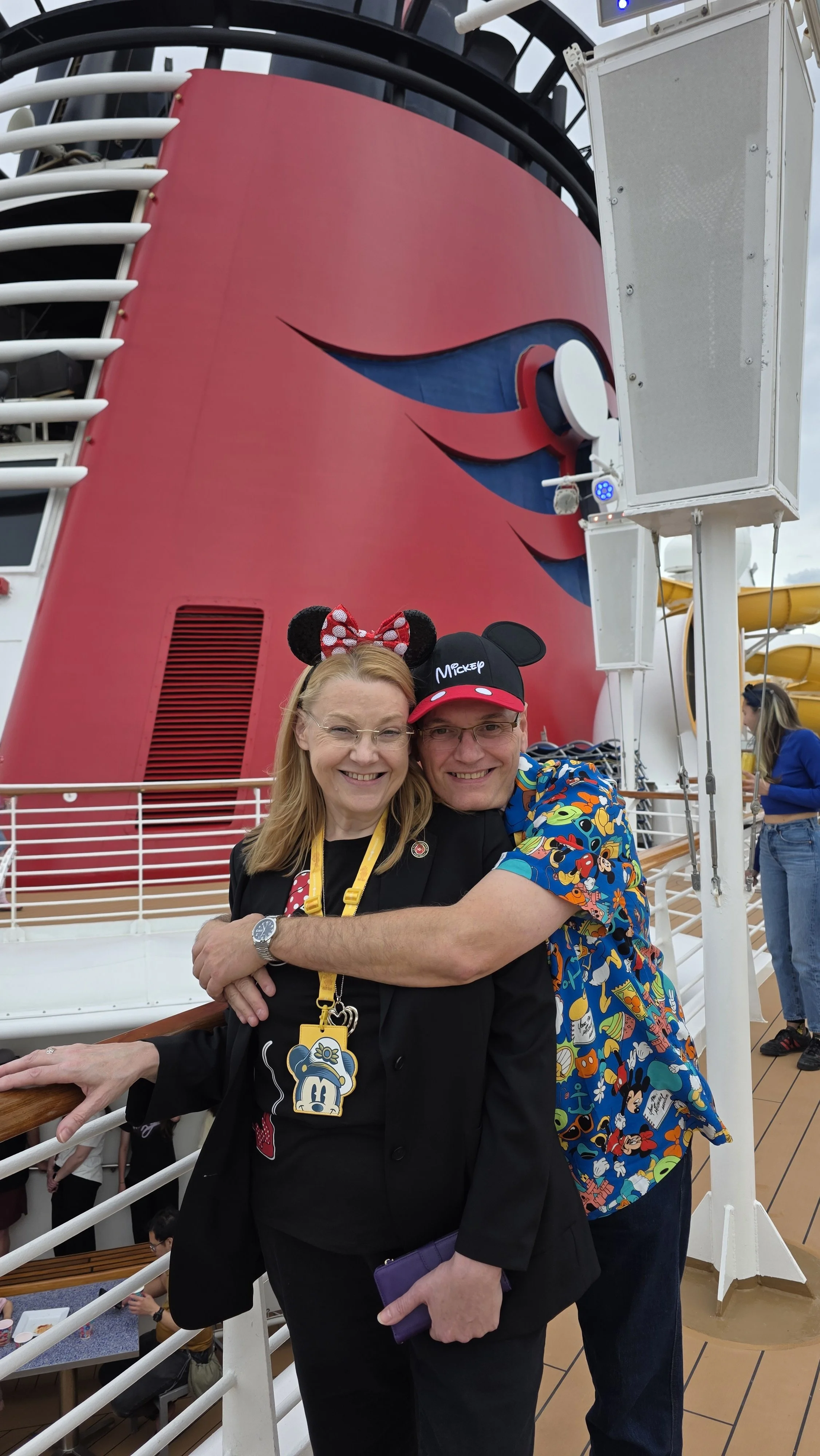 Smiling woman and man wearing Mickey Mouse-themed accessories, hugging on the deck of a cruise ship with a large red and black ship structure in the background.