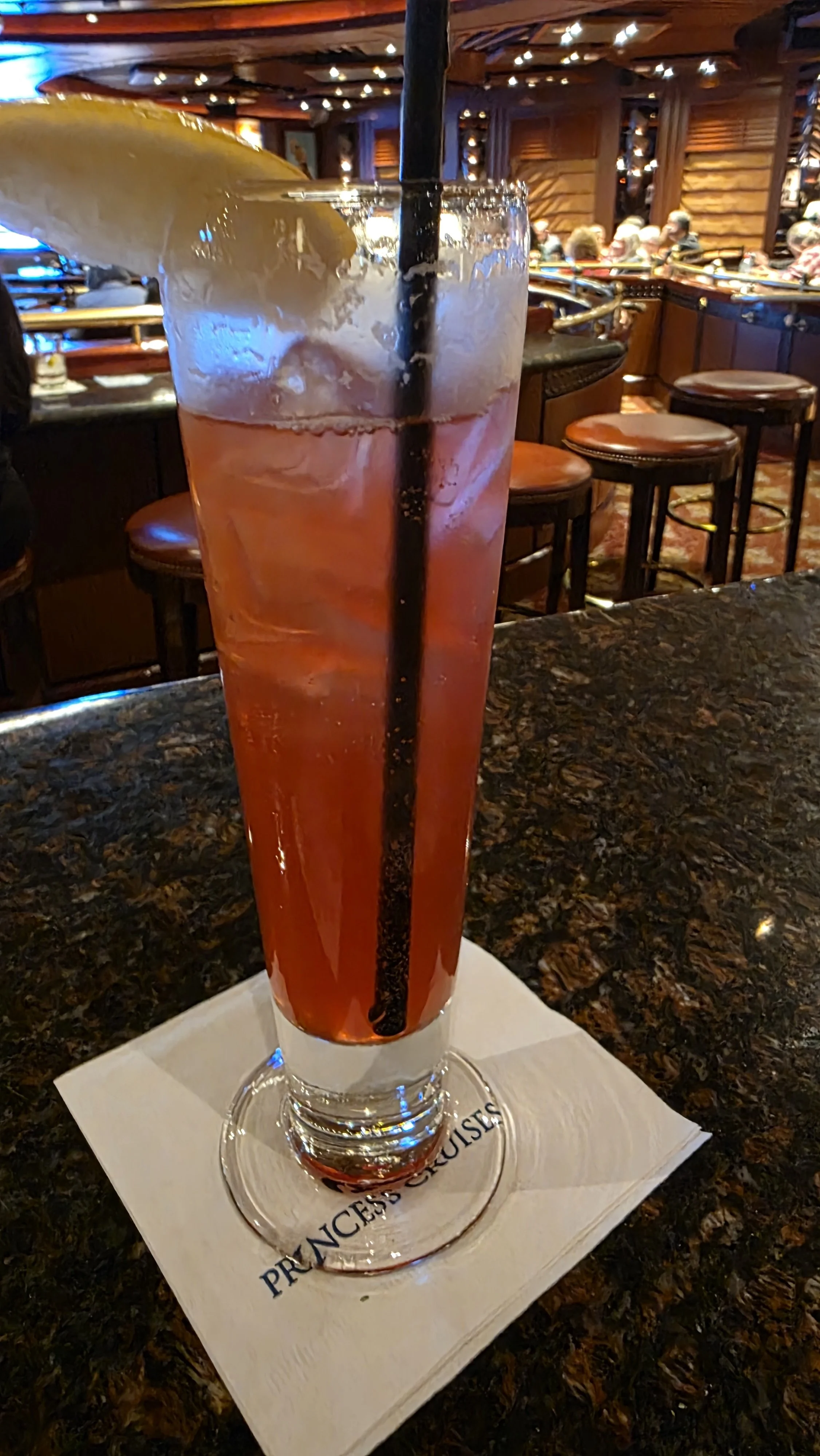 A tall glass of pink-colored cocktail with a lemon wedge garnish and a black straw, placed on a napkin on a bar counter in a dimly lit, rustic-themed restaurant or bar with wooden walls and stools.