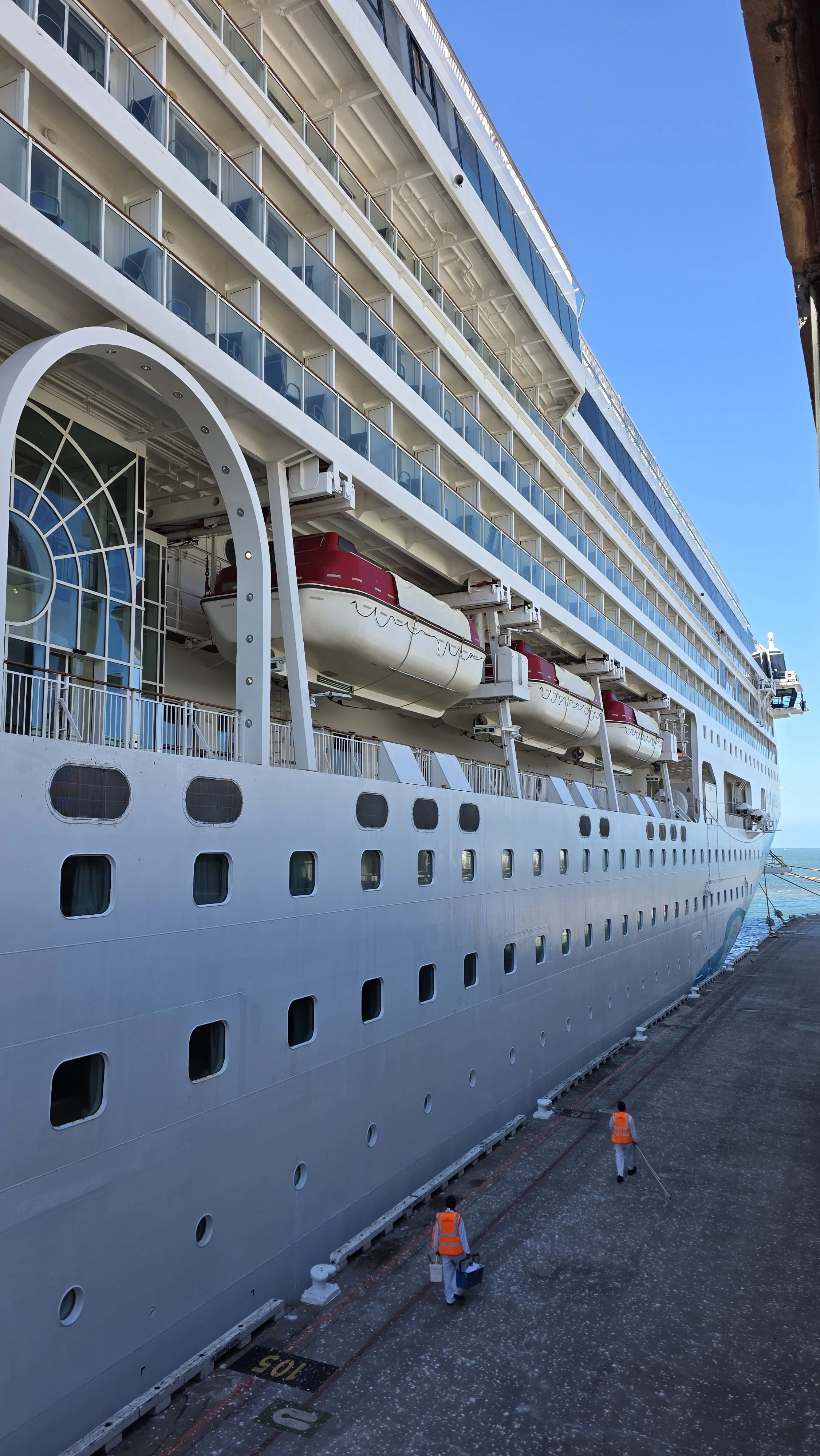 A large white cruise ship docked at a port, with workers in orange vests walking on the dock. The ship has multiple decks with balconies and lifeboats visible along the side.