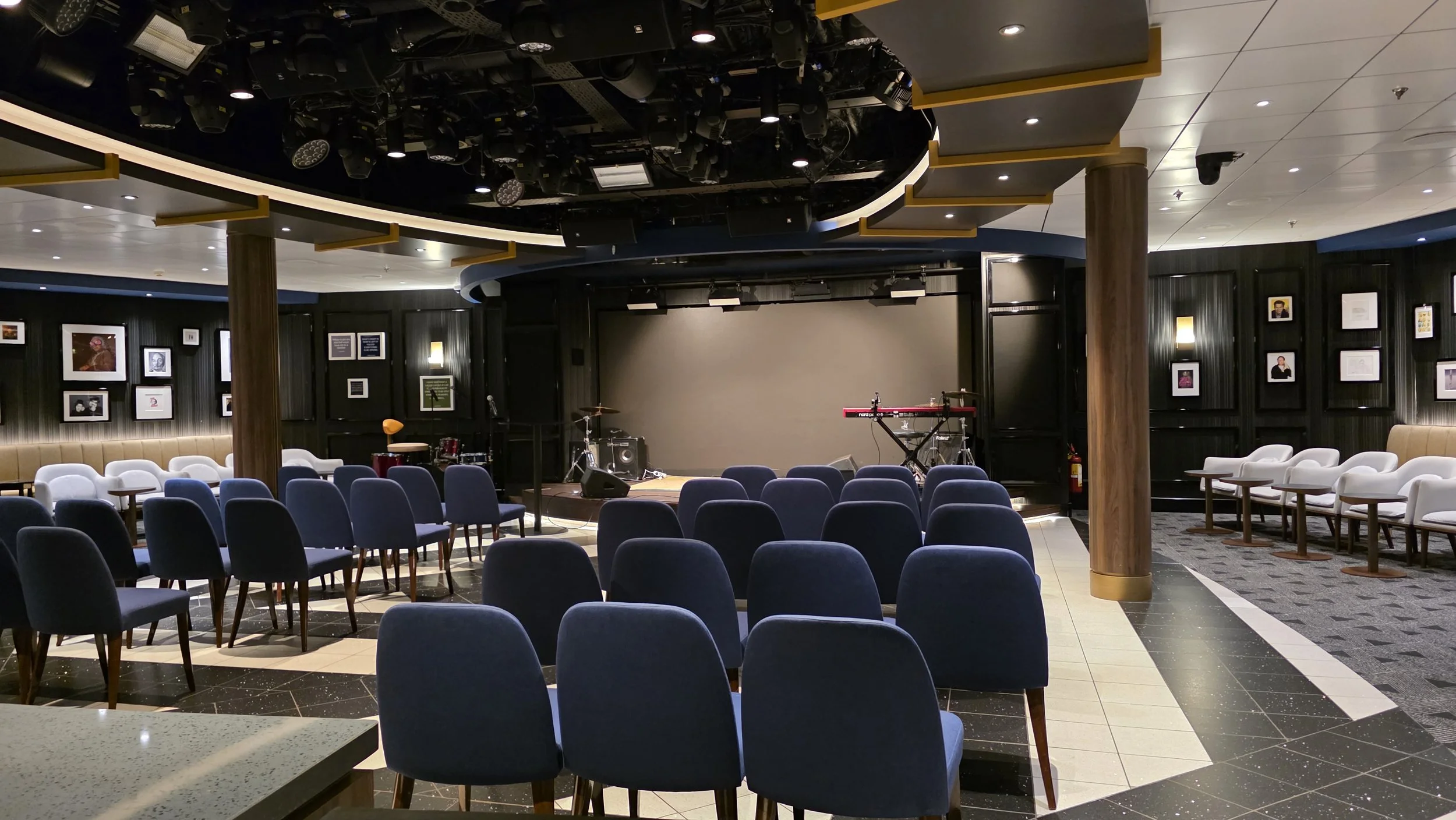 Indoor performance venue with a small stage, musical instruments, chairs arranged for an audience, and framed photos on the black walls.