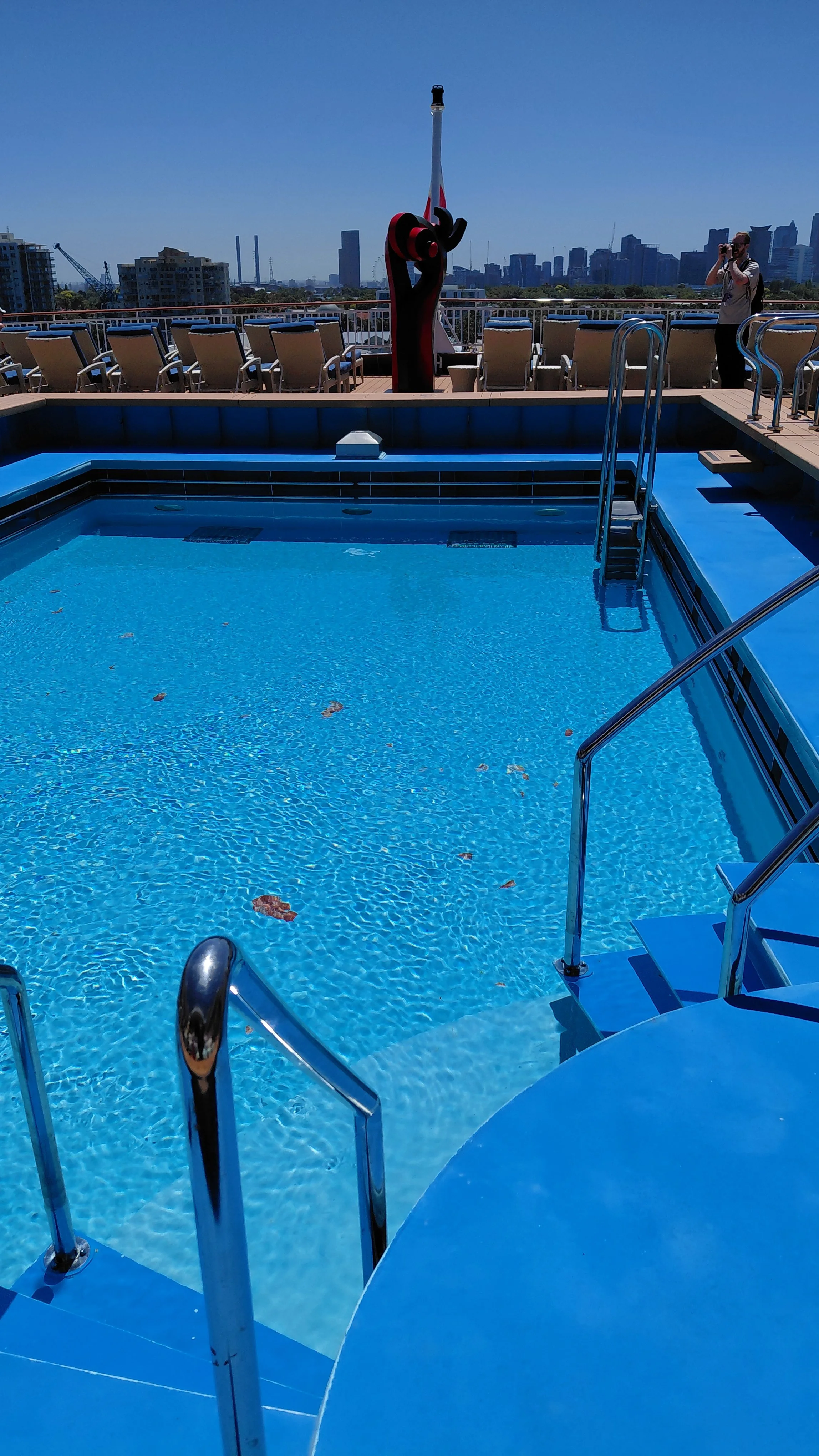 Empty rooftop swimming pool with blue water, metal handrails, and city skyline in the background. A person is taking a photo on the deck near the pool, and another sculpture is on the deck.