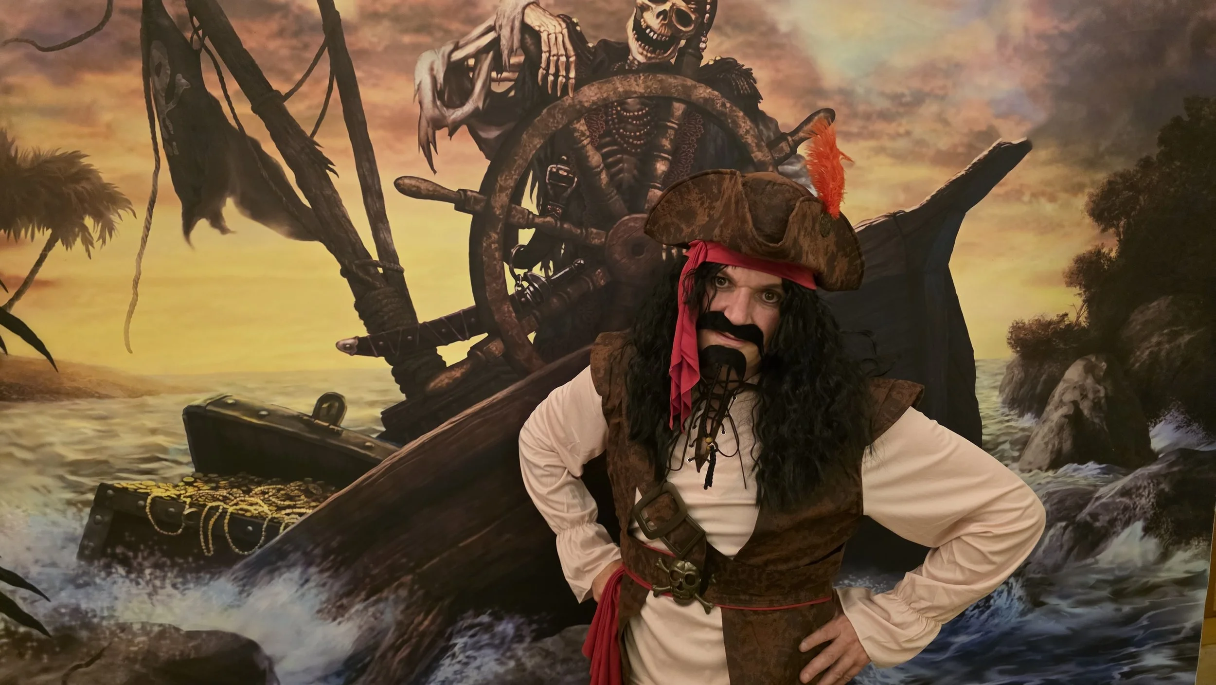 Person dressed as a pirate with a large hat and beard standing in front of a pirate ship scene with a sunset background.