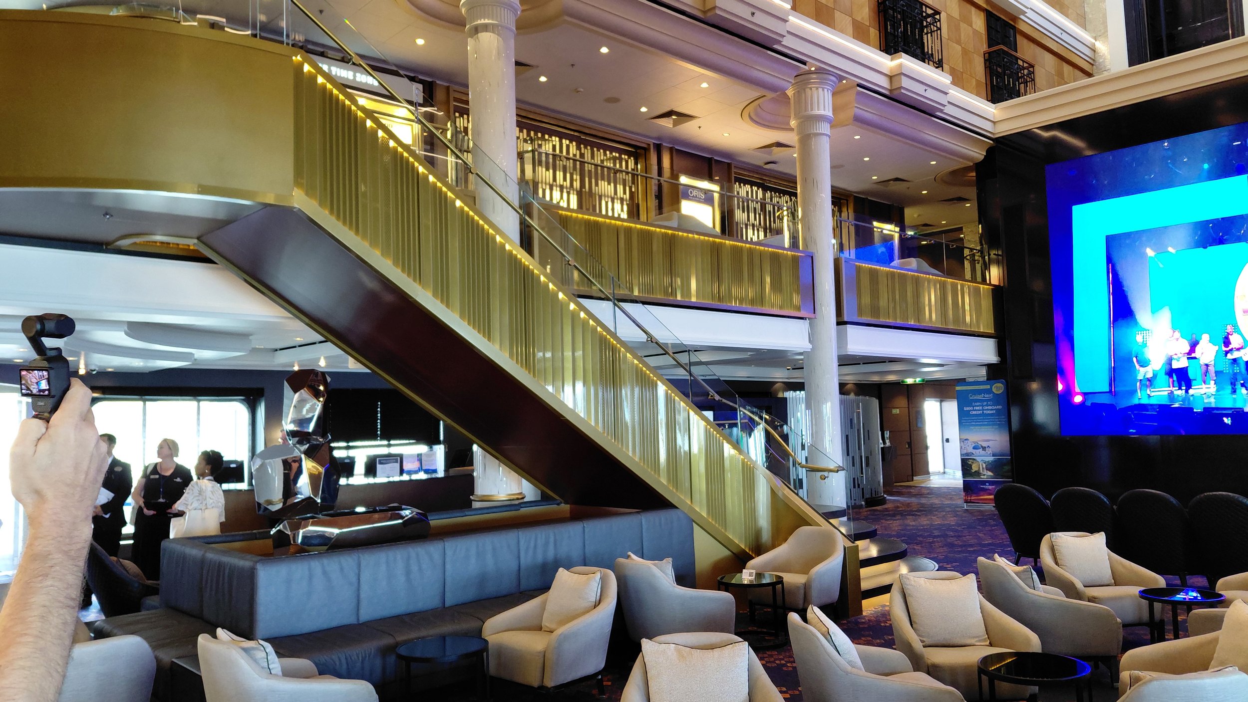 Interior of a luxurious cruise ship lounge with seating, a large screen displaying a performance, a prominent gold staircase, and decorative sculptures.