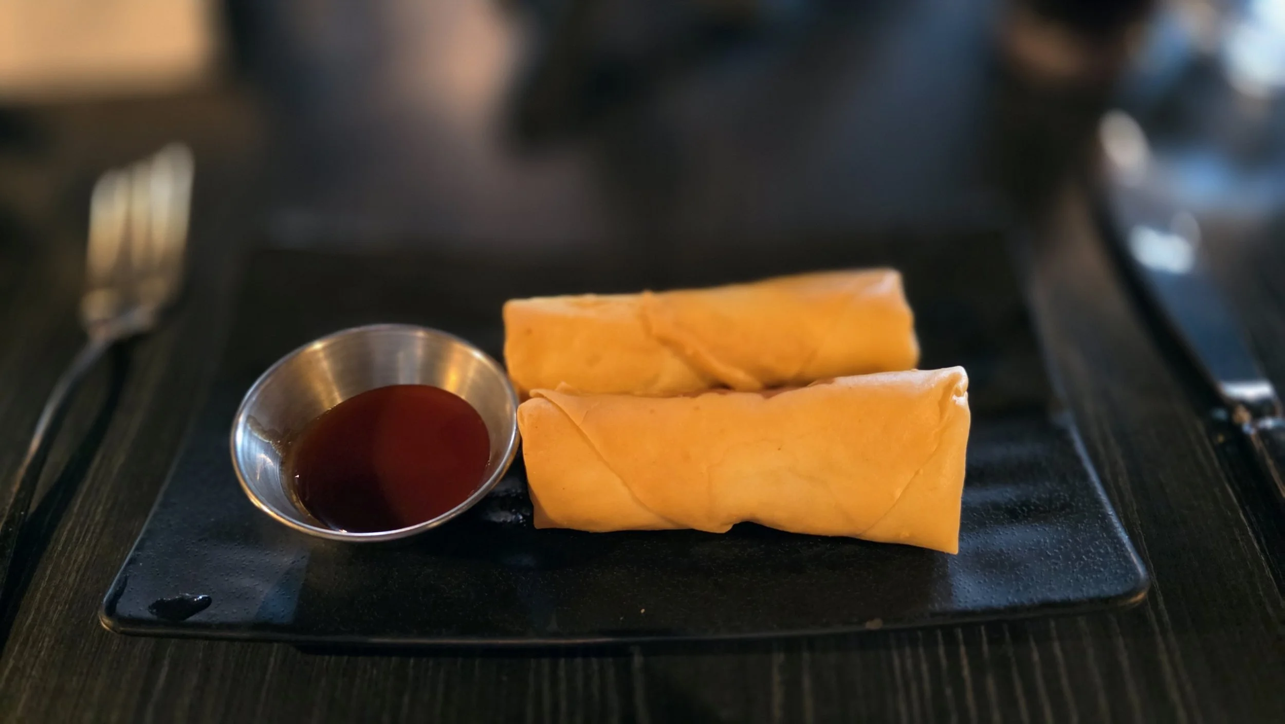 Two spring rolls on a black plate with a small metal cup of dipping sauce, likely soy or sweet chili, on the side.