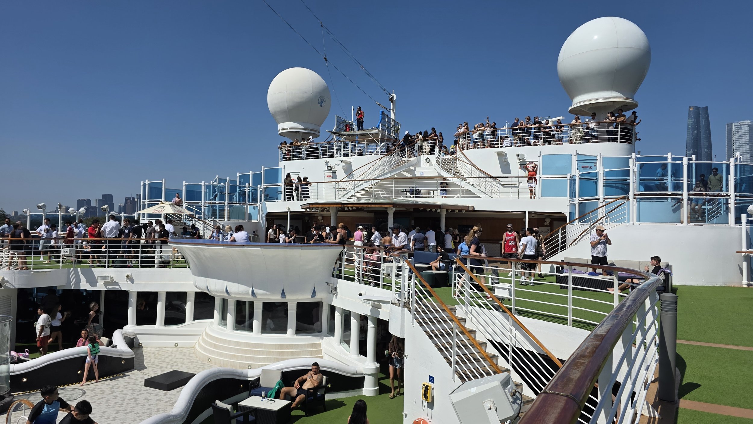 Crowded upper deck of a cruise ship with people enjoying the sunny weather, two large radar domes, and a city skyline in the background.