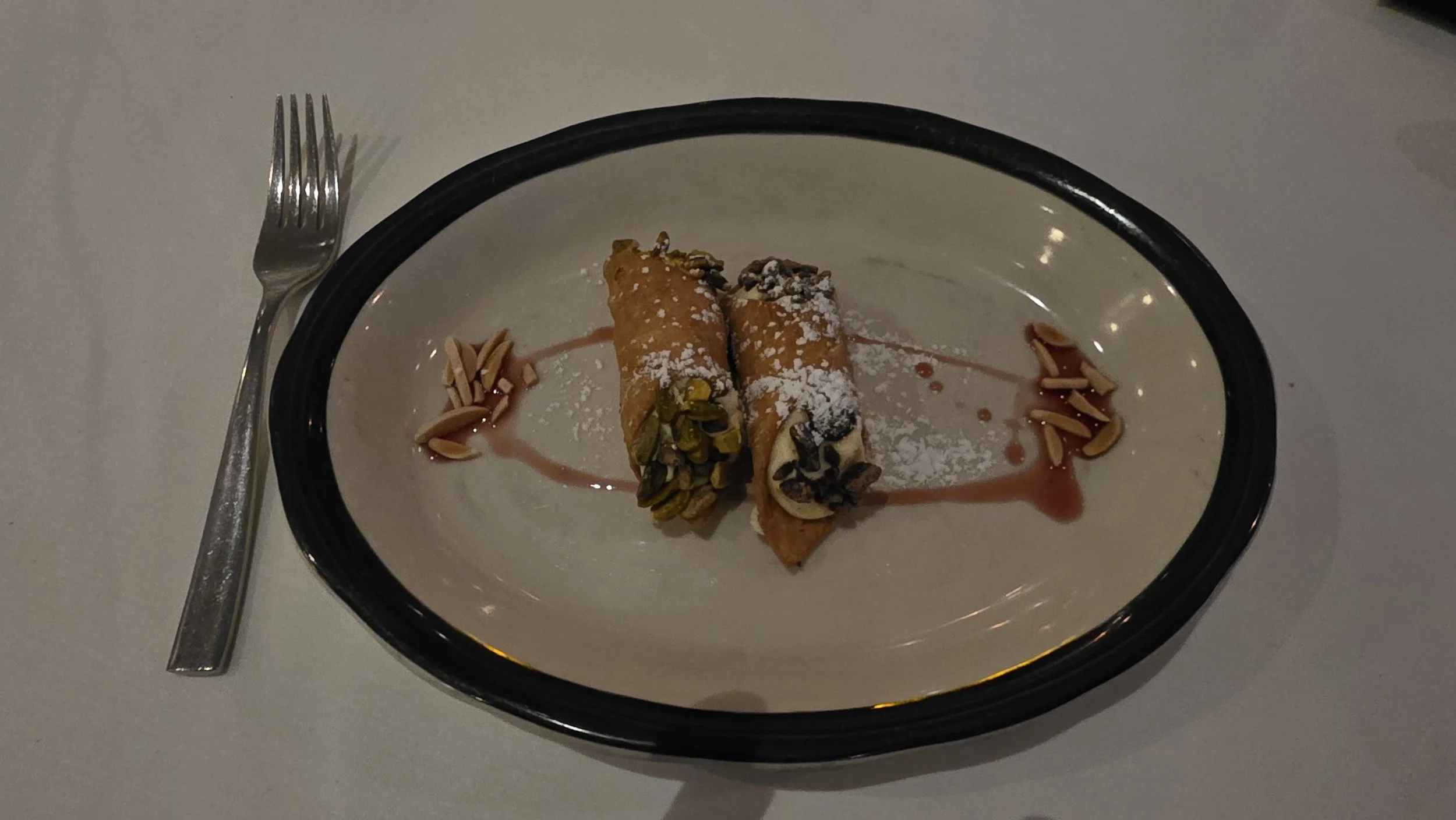 Two cannoli desserts garnished with pistachios, powdered sugar, and a drizzle of sauce, served on a white plate with black rim, accompanied by a fork on a white table.