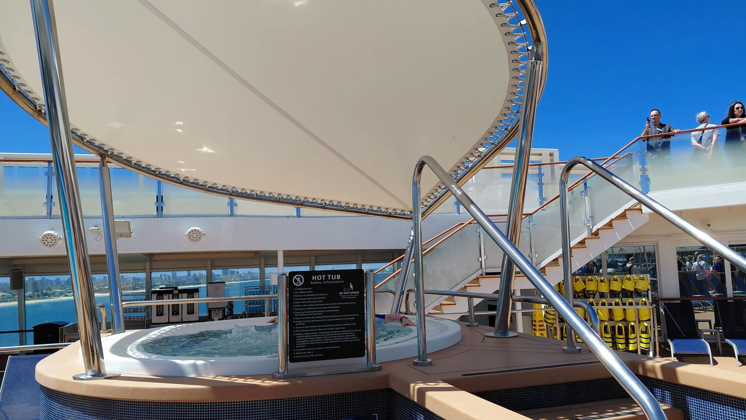 A hot tub on a cruise ship deck, with a group of people visible in the background. The hot tub is covered by a large white canopy, and there are safety instructions displayed on a sign in front of it. The scene shows clear blue skies and a distant ci
