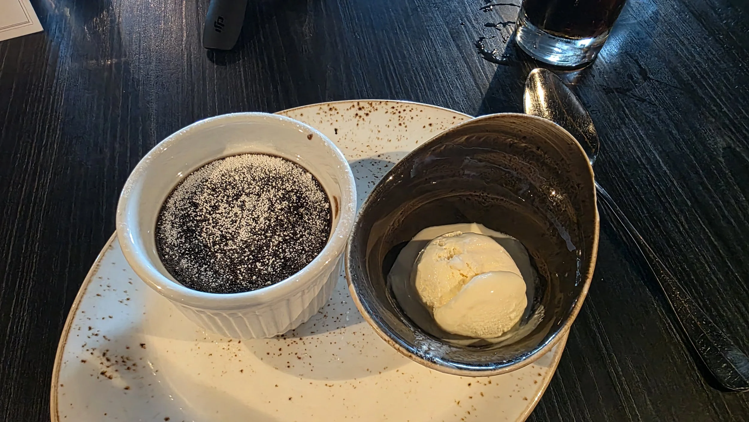 A dessert plate with a small ramekin of chocolate pudding topped with powdered sugar, a bowl of vanilla ice cream, a spoon, and a glass of water on a dark wooden table.