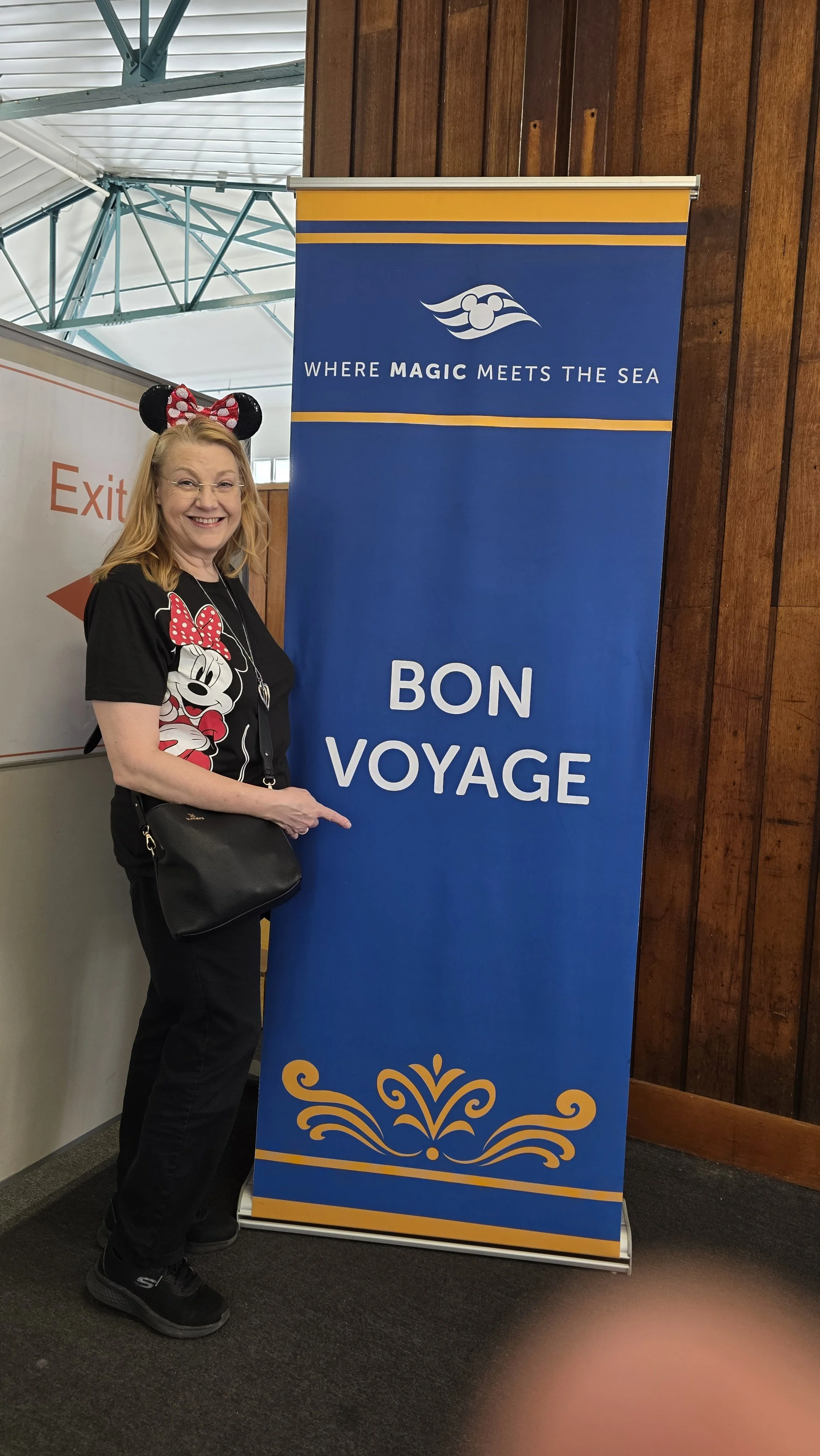 A woman wearing Mickey Mouse themed clothing and accessories standing next to a blue banner that says 'Bon Voyage' at an airport or terminal.