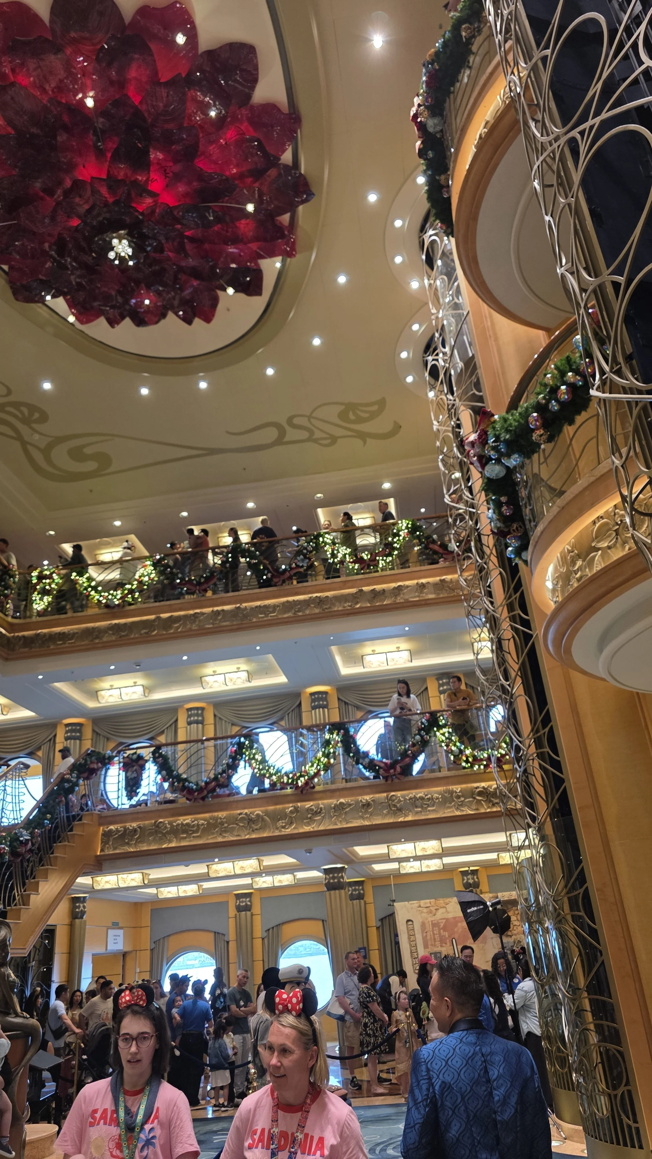 Indoor lobby decorated with holiday garlands, balloons, and large red floral ceiling installation, crowded with people including two women wearing Minnie Mouse ears, some in line or waiting.