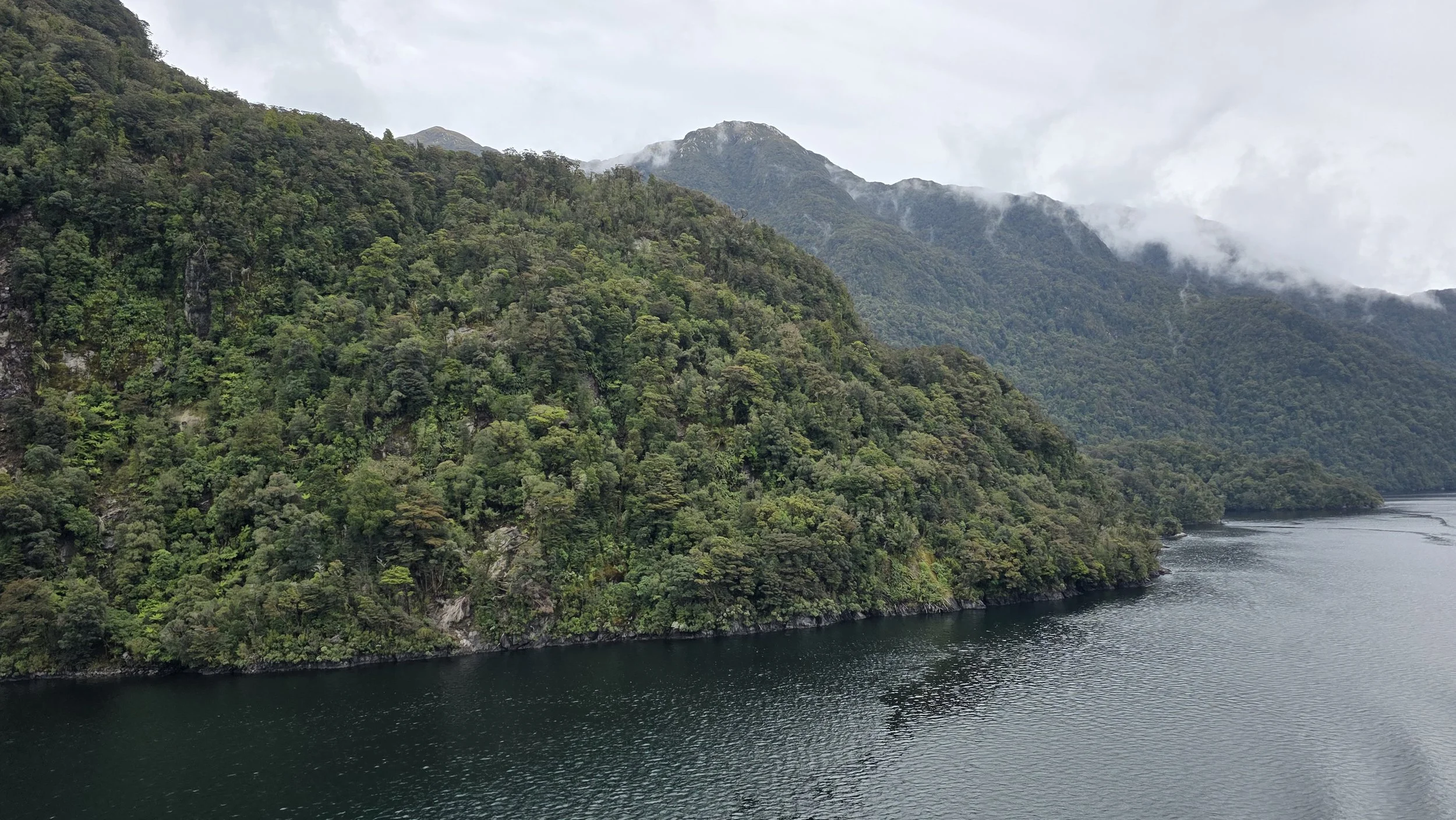A landscape of lush green forested mountains with a body of water at the base, and mist or clouds partially covering the mountain peaks.