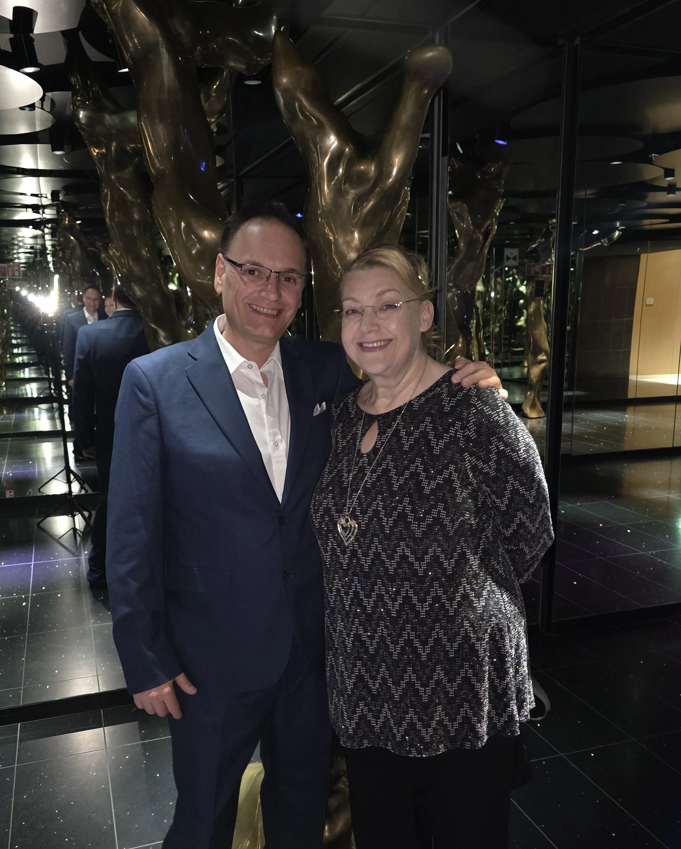 A man and woman are smiling and posing together in front of a decorative golden sculpture and mirrored walls.