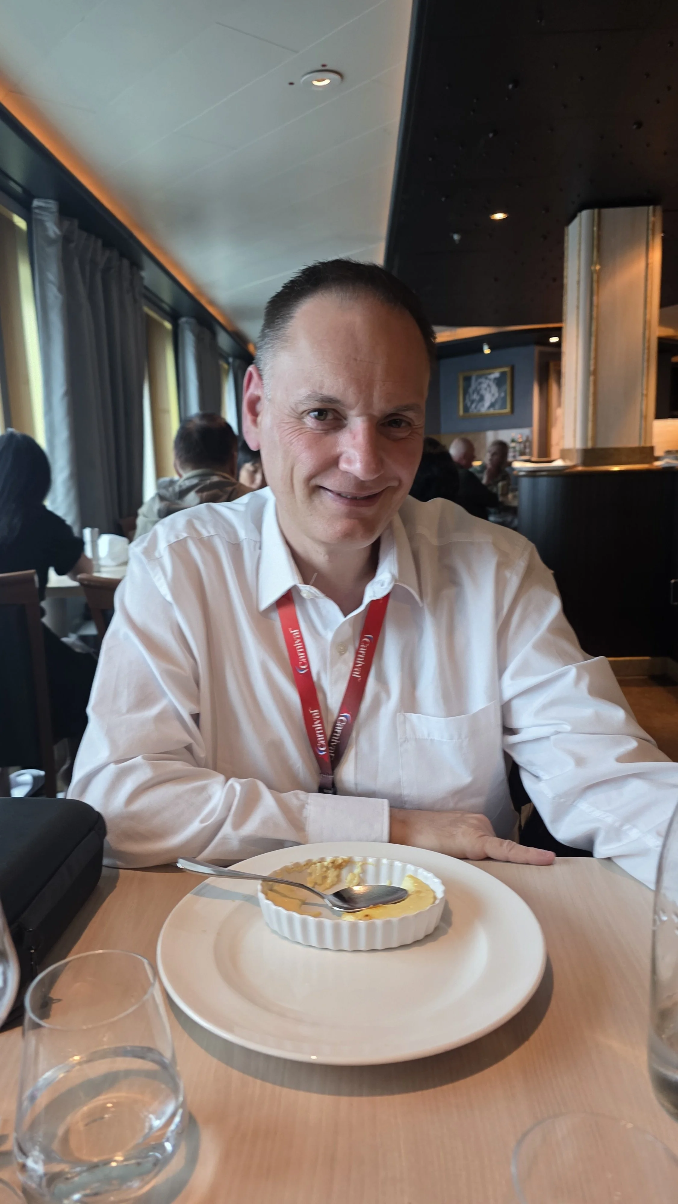 A man in a white shirt sitting at a table in a restaurant with a small dish and spoon in front of him, smiling at the camera.