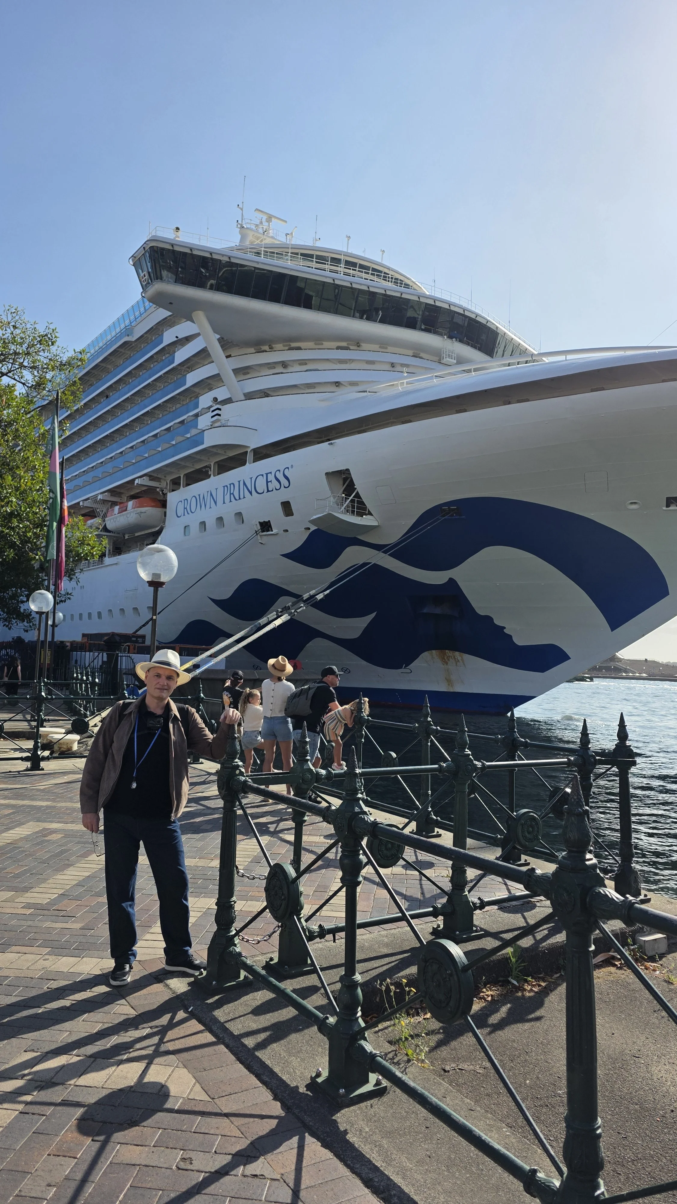 Large cruise ship named Crown Princess docked at the port with people standing nearby, some taking photos. A man holding a leash is in the foreground, and the sky is clear.