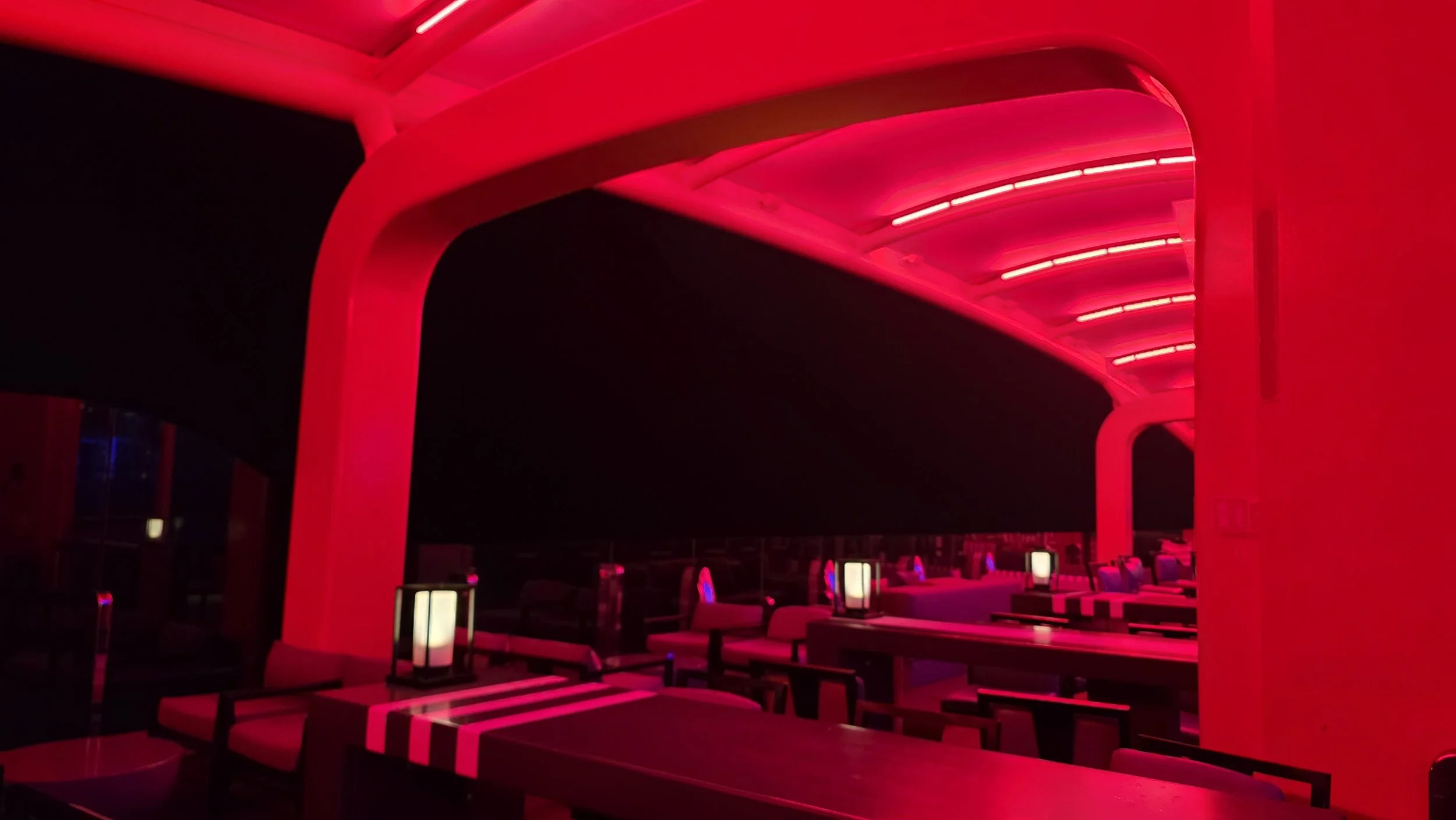 Night view of a modern, covered outdoor seating area illuminated by pink and purple lights, with chairs, tables, and lit lamp posts.