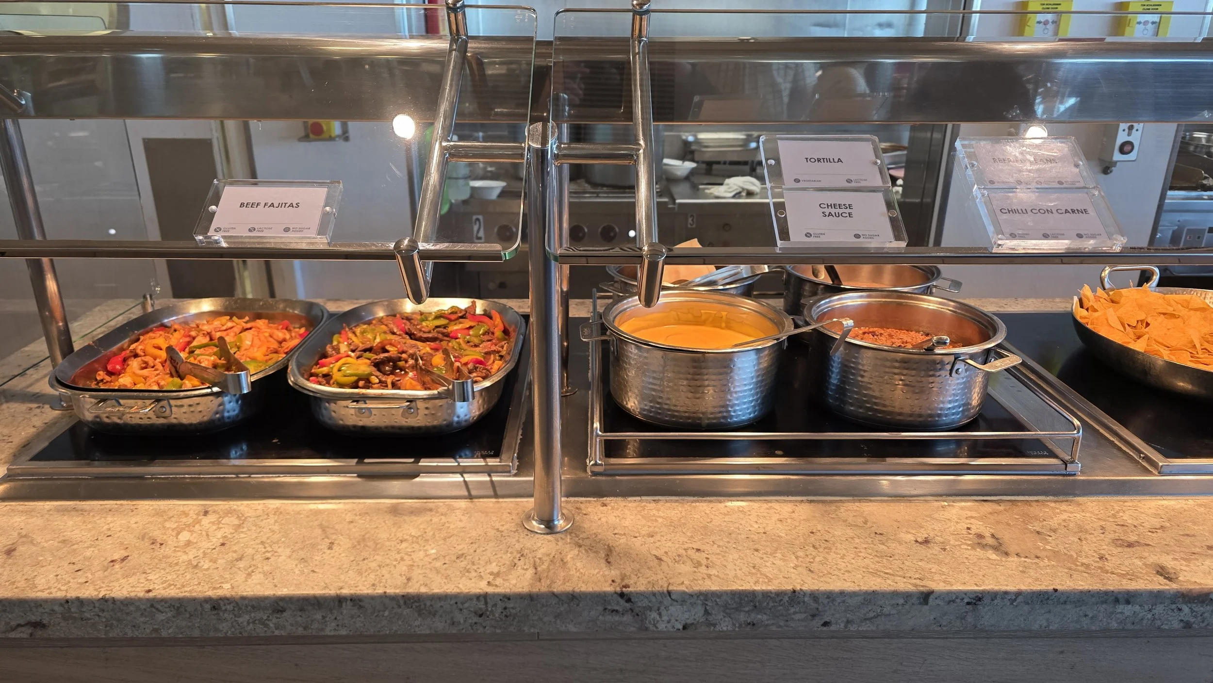 Buffet station with trays of food including beef fajitas, sliced peppers, cheese sauce, chili con carne, and tortilla chips, with labels.