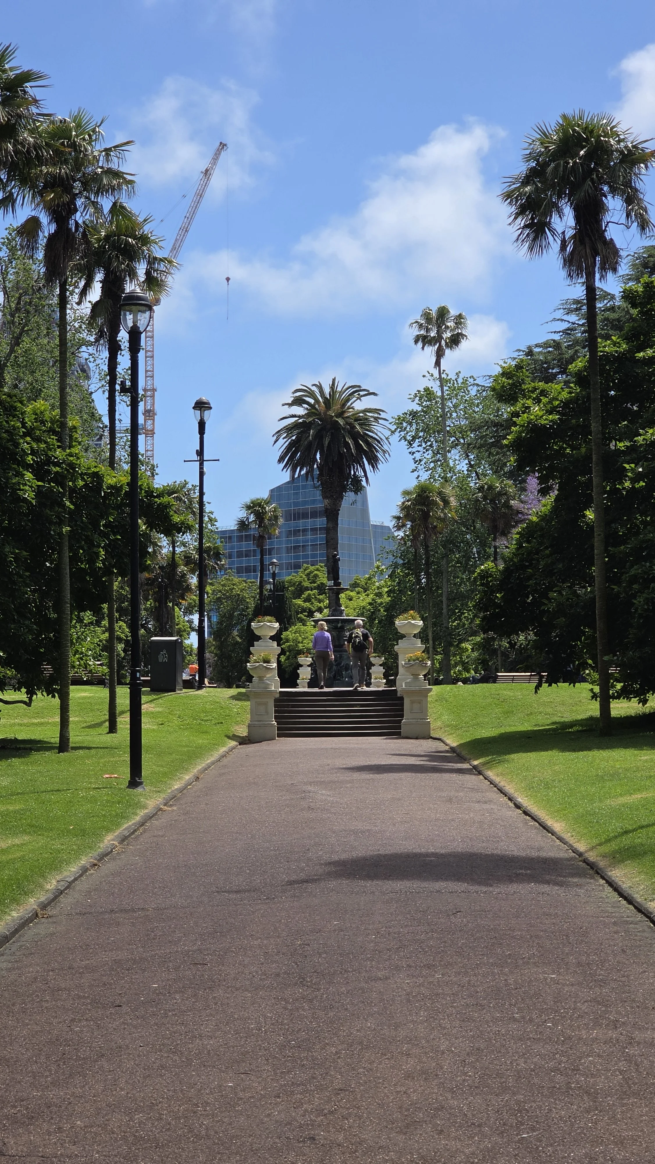 Park pathway leading to a monument with palm trees and a modern glass building in the background under a partly cloudy sky.