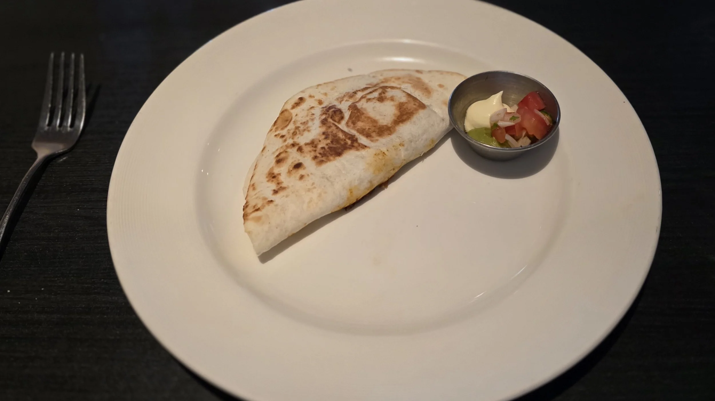 Plate with a folded quesadilla and a small bowl of salsa with sour cream, set on a dark table, with a fork to the left.