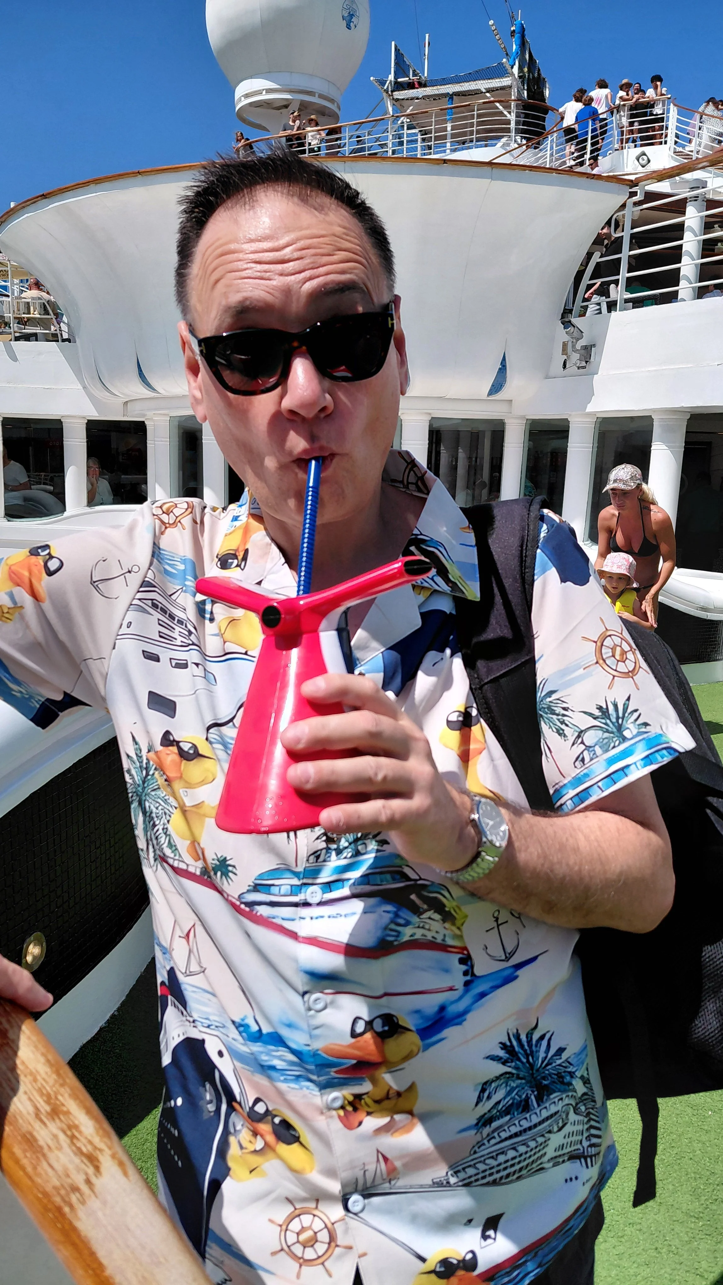 A man wearing sunglasses and a colorful tropical shirt is sipping a beverage through a pink and blue straw on a cruise ship. The background shows the ship's deck with other passengers, a white exterior, and a blue sky.