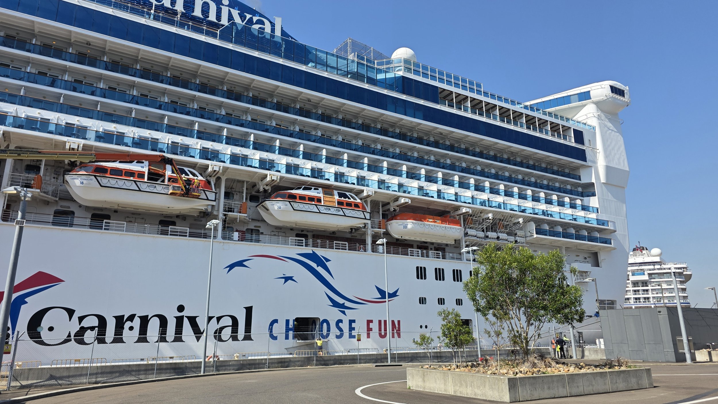 The Carnival cruise ship docked at a port with clear blue skies overhead. The ship's side displays the word 'Carnival' and colorful graphics, including the slogan 'Choose Fun.' Several lifeboats are visible on the side of the ship, and there are some