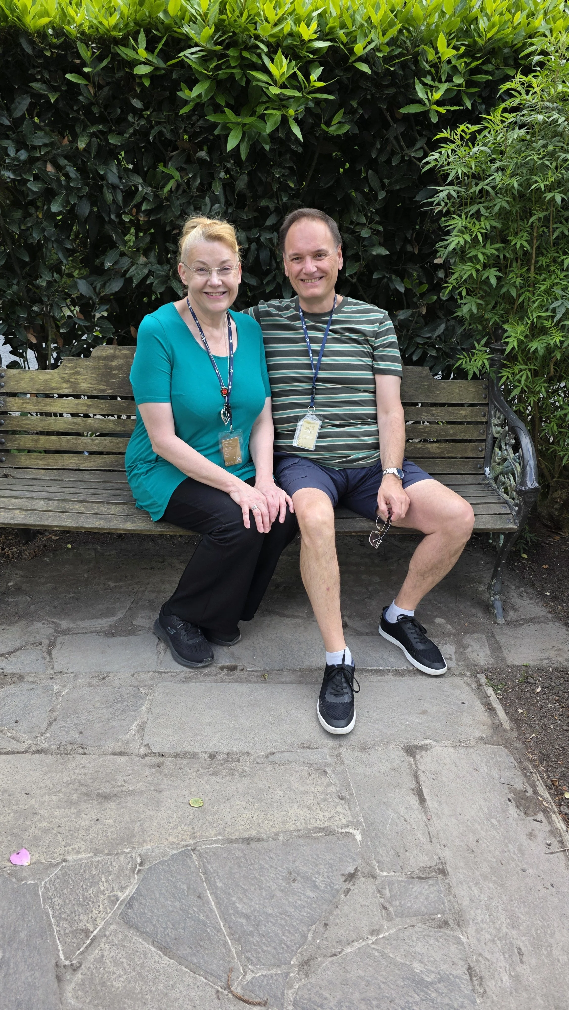 Two people, an older woman and a younger man, sitting on a wooden park bench outdoors, smiling. The woman wears a teal blouse and black pants, and the man wears a striped t-shirt and shorts. They have lanyards with badges around their necks, and ther