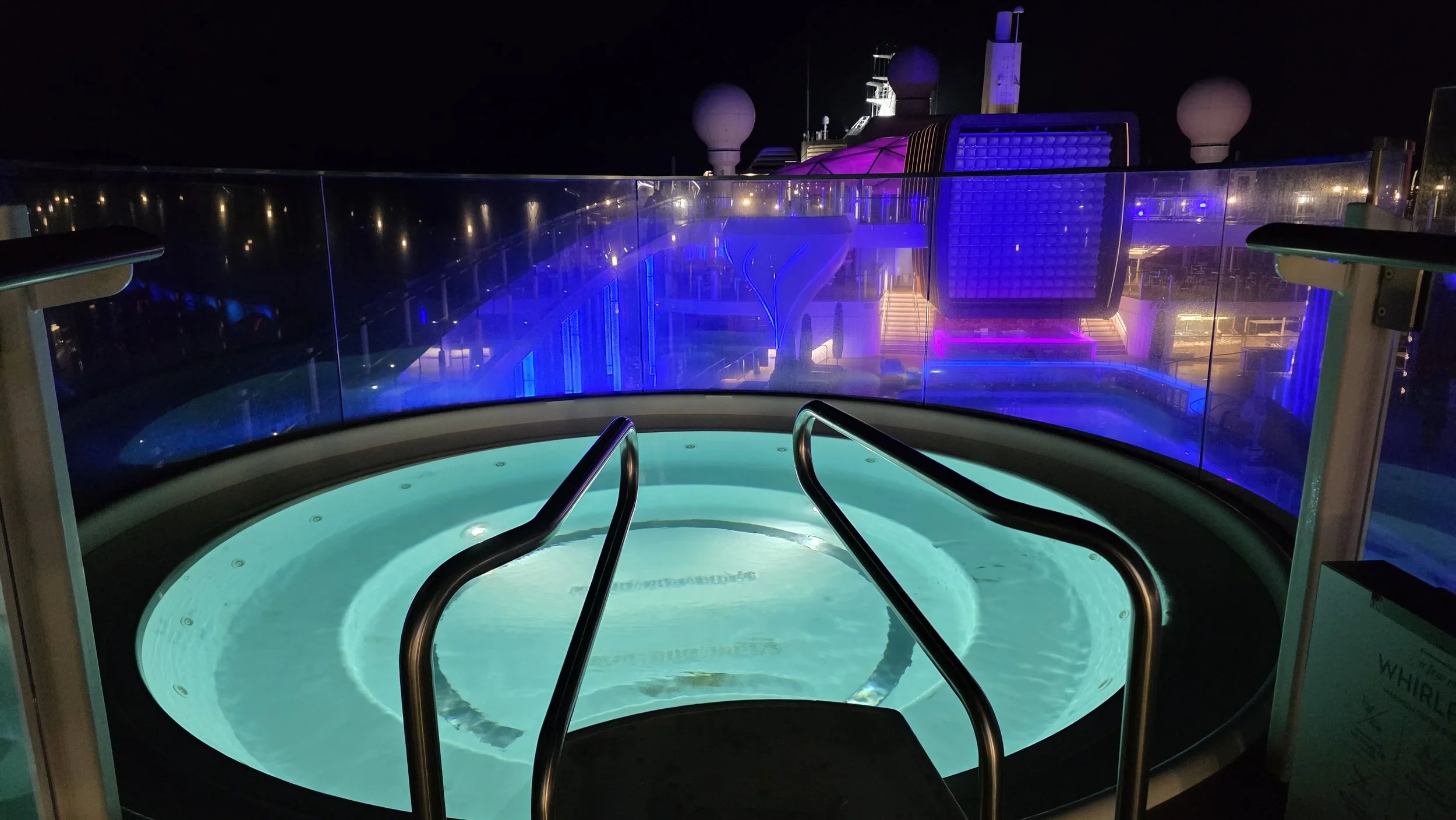 A glowing hot tub on a cruise ship deck at night with city lights in the distance, illuminated by blue and purple lights.