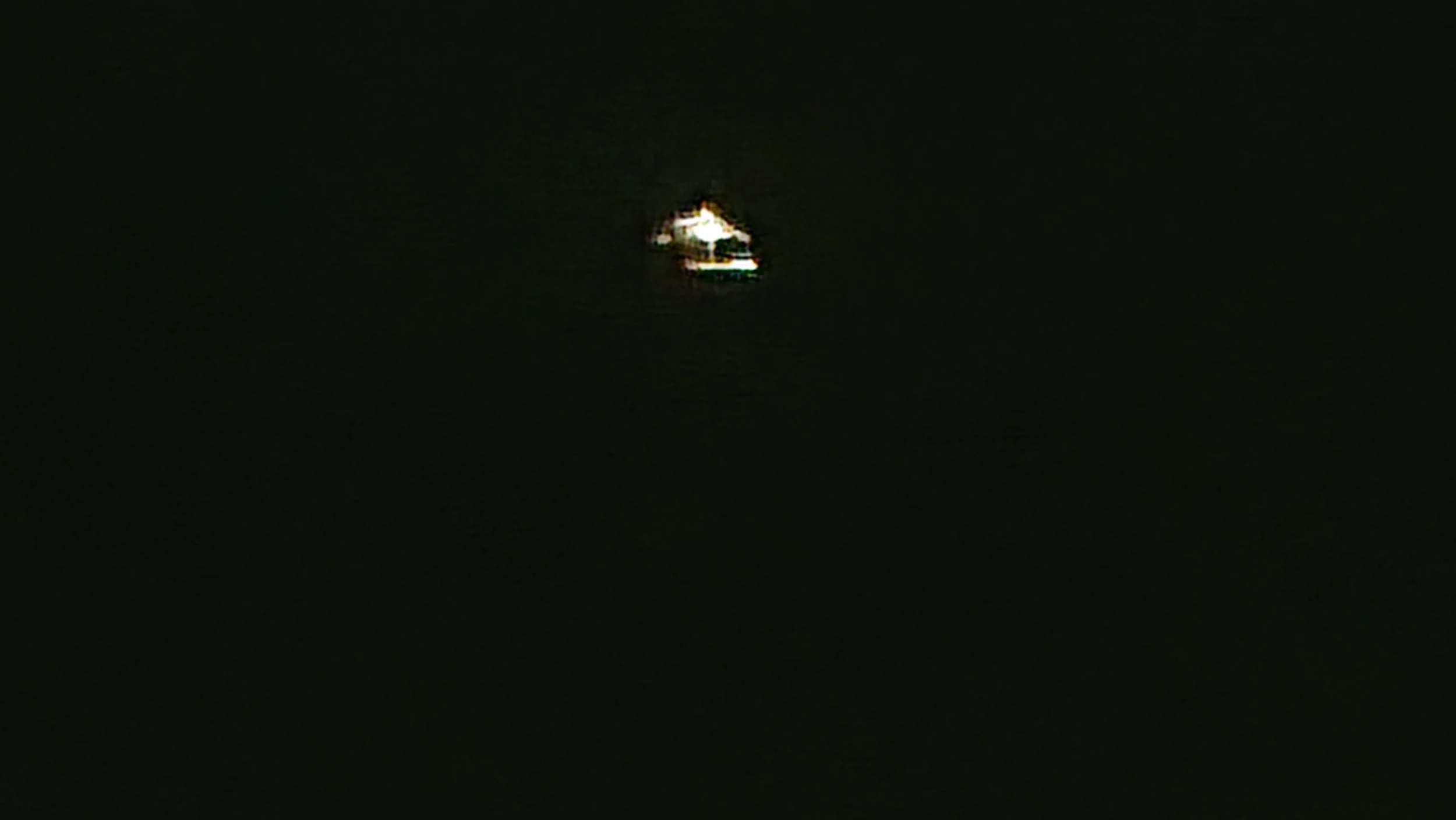 A small boat with white and green lighting reflects on dark, calm water at night.