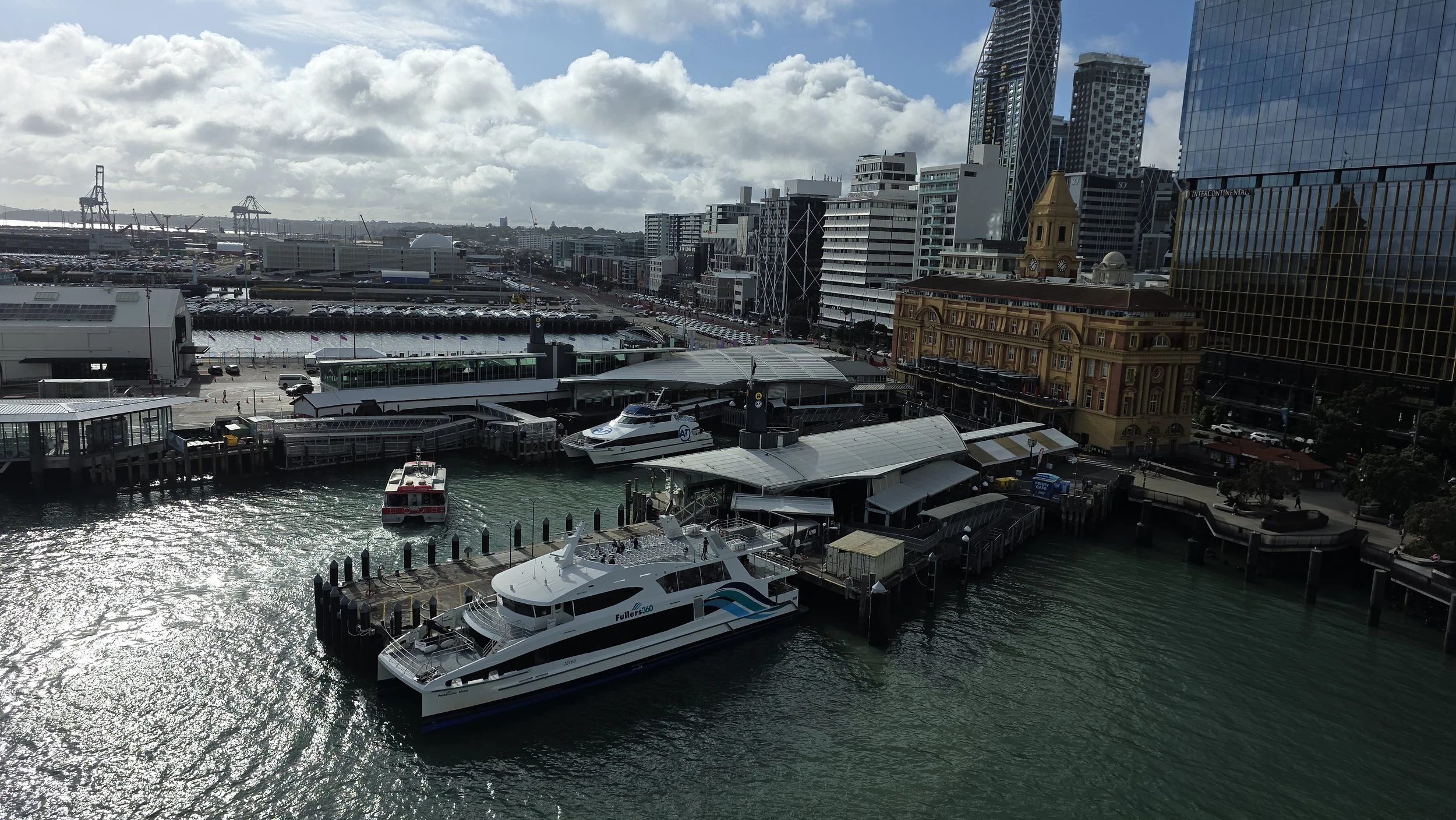 Cityscape of a harbor with boats docked, including a large white yacht, and tall modern buildings in the background under partly cloudy skies.