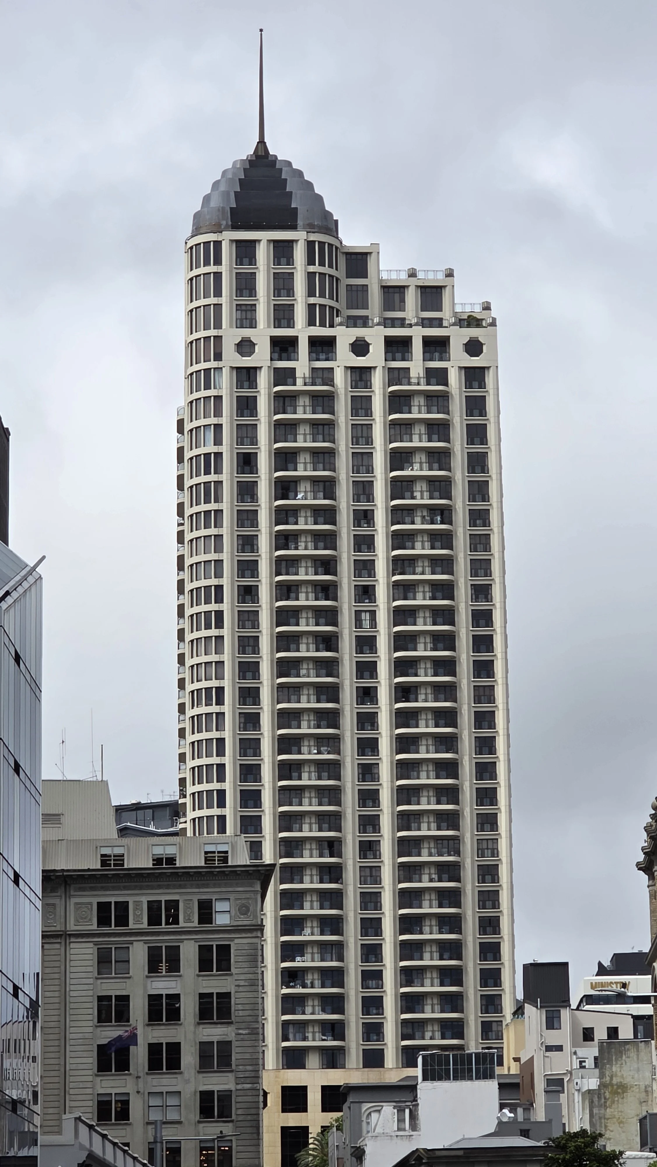 Tall modern skyscraper with rounded balconies and a pointed tip at the top, surrounded by other buildings in an urban cityscape under a cloudy sky.