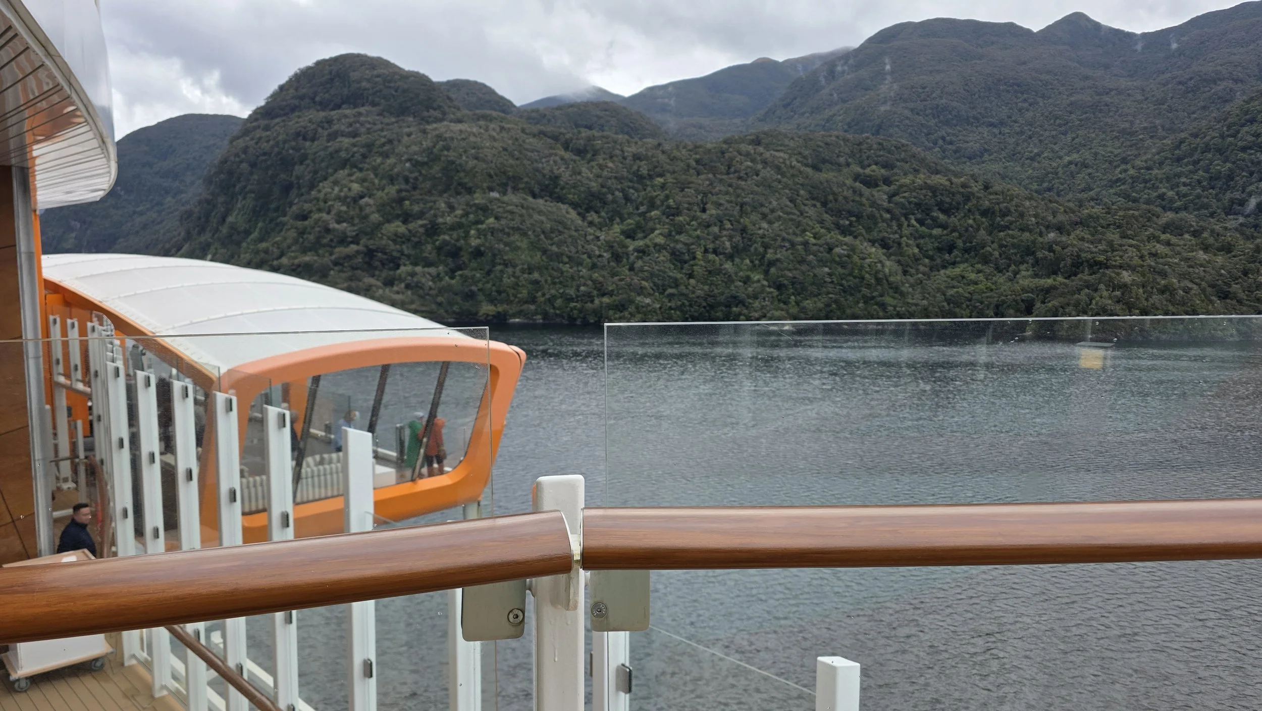 View from a cruise ship balcony overlooking a lake and green mountains under a cloudy sky.