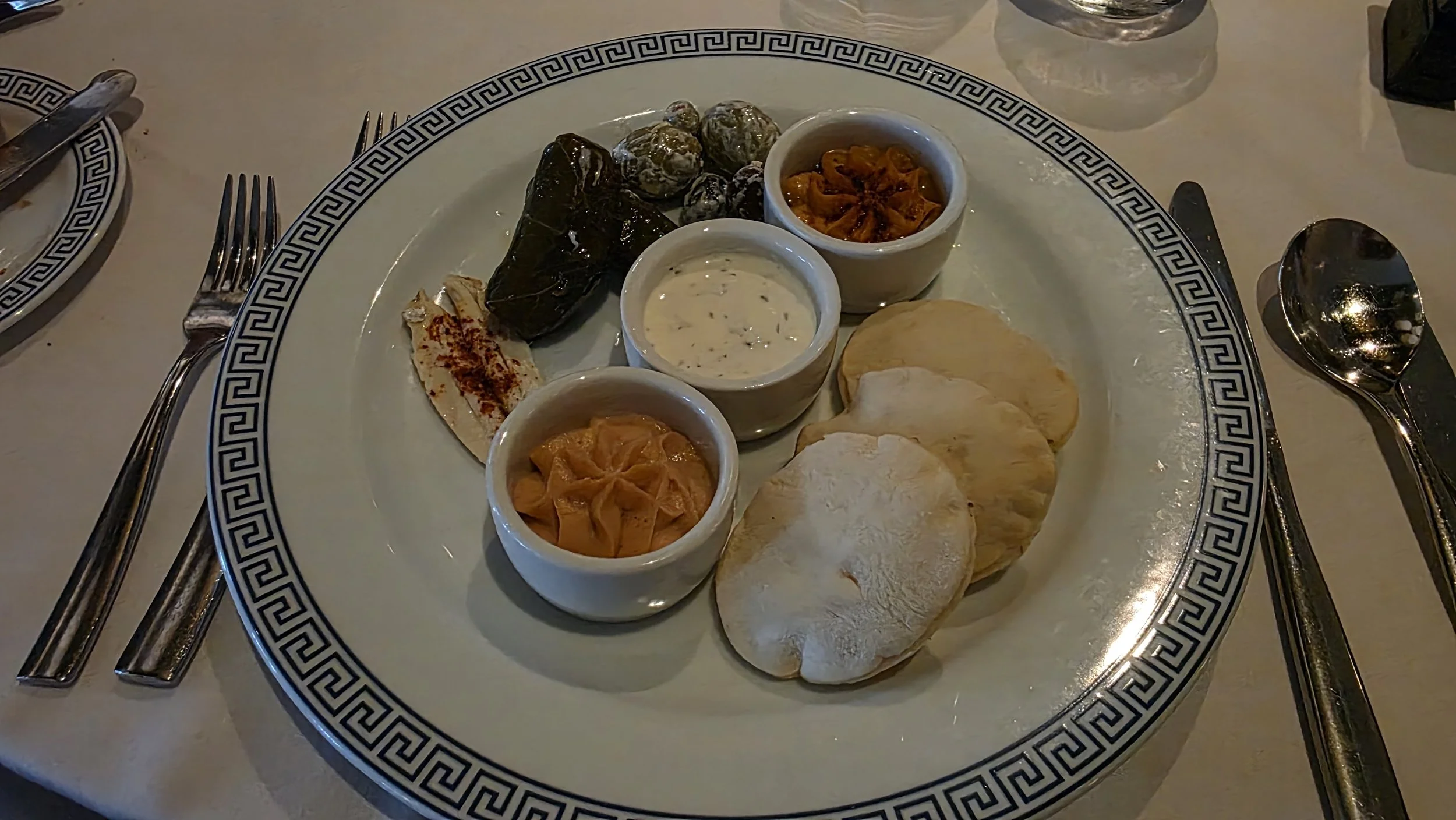 A large plate with various Middle Eastern appetizers and dips including olives, roasted peppers, hummus, and pita bread, set on a dining table with utensils and glasses around.